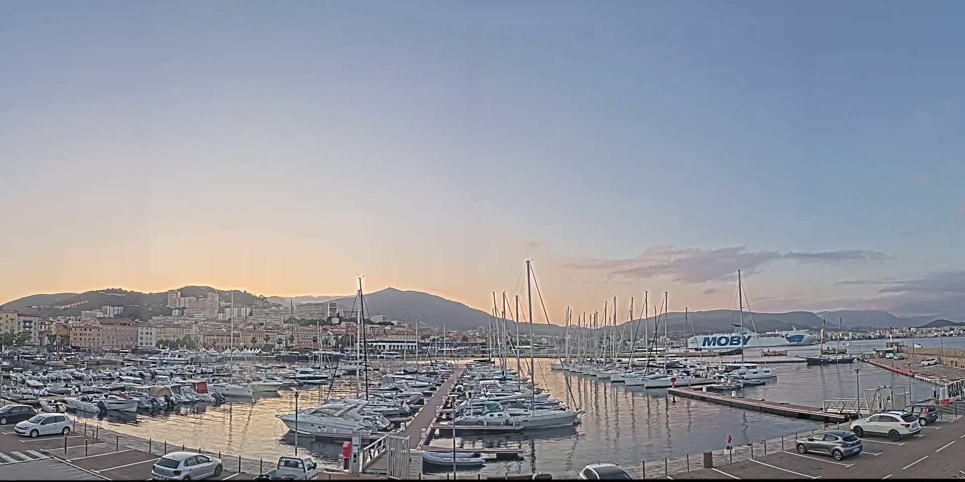 A marina filled with numerous boats is situated in front of a city nestled against mountains under a clear, twilight sky.