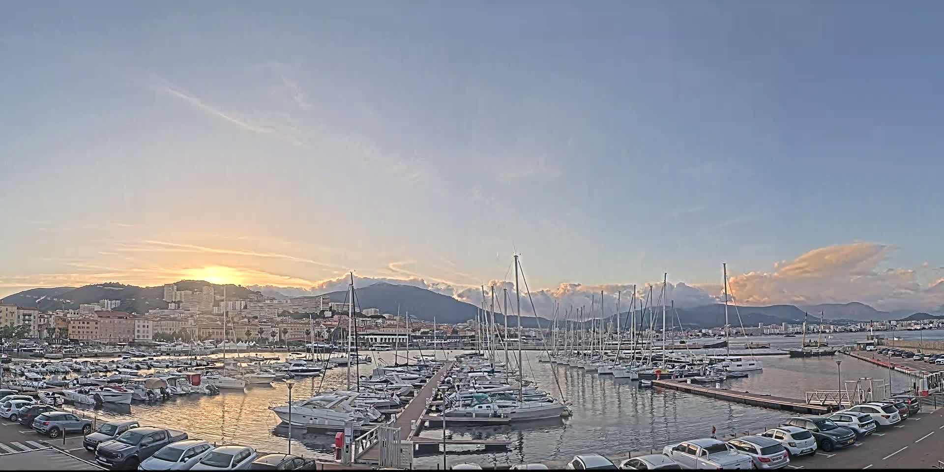 A harbor filled with sailboats and yachts is situated in front of a city at sunset under a partly cloudy sky.