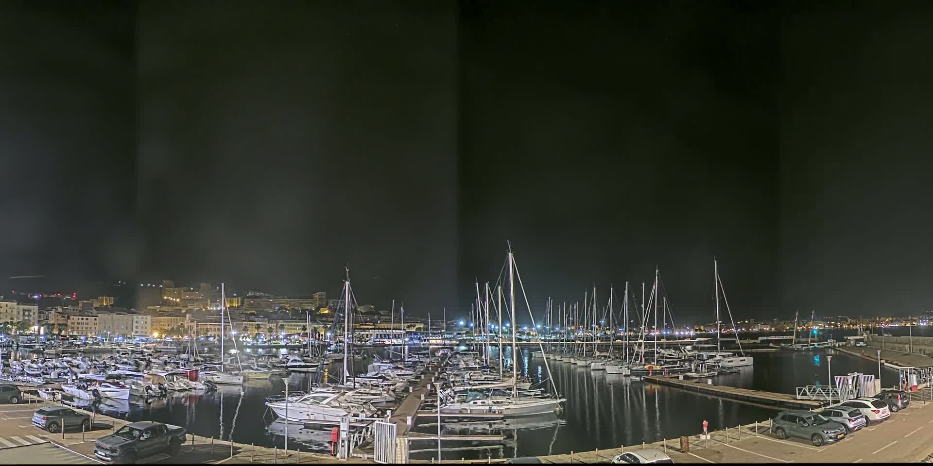 A marina filled with sailboats and motorboats at night, under a dark sky, is illuminated by lights on the docks and buildings in the background.