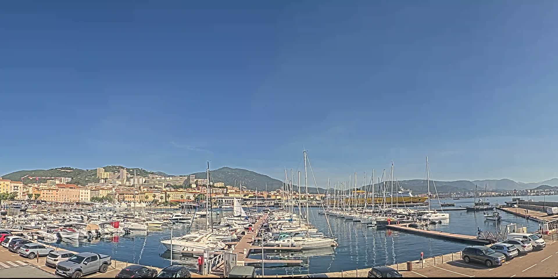 A marina filled with numerous sailboats and motorboats is situated in front of a city nestled against a mountain range under a clear blue sky.