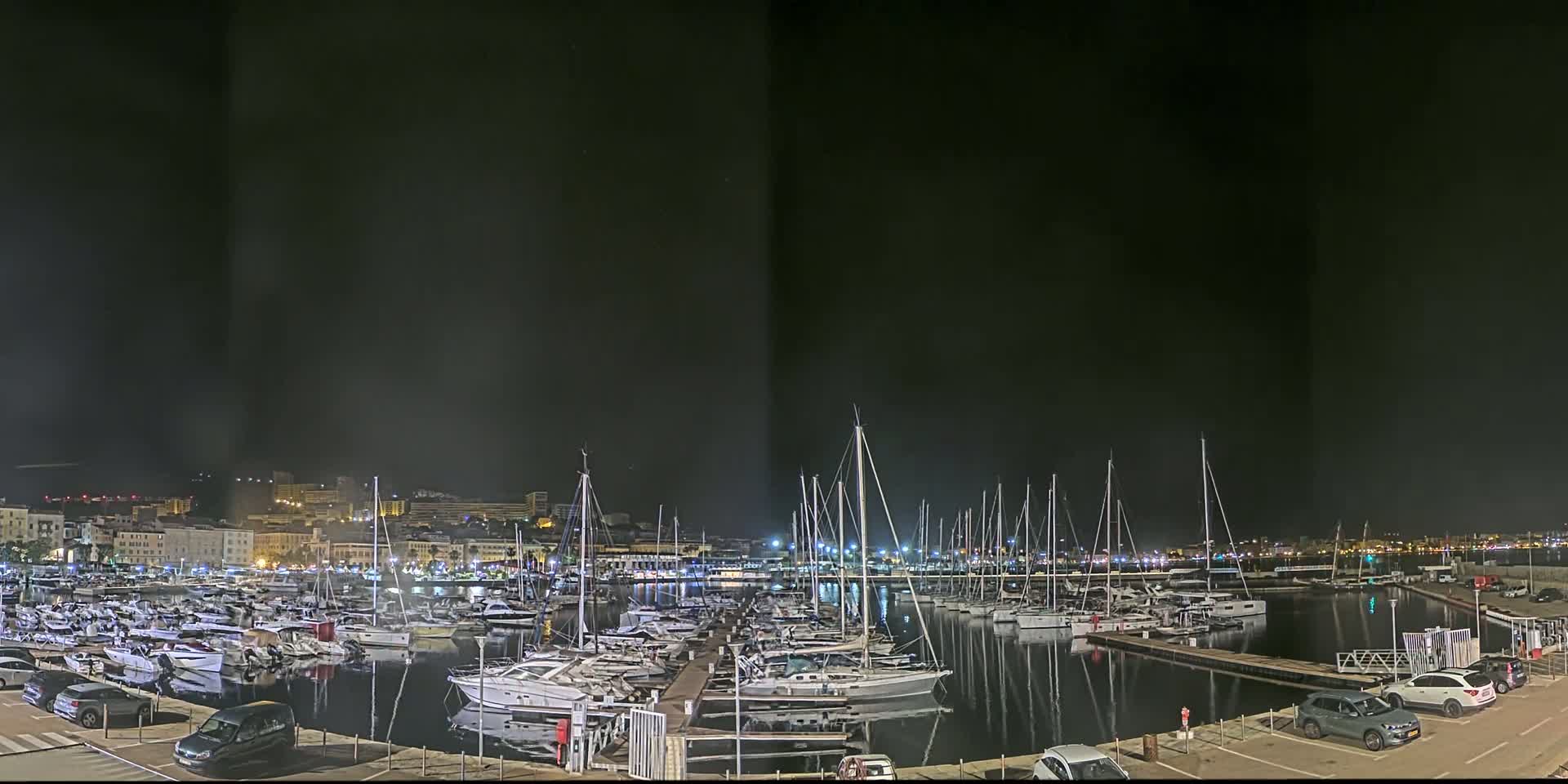 A marina filled with numerous boats and yachts is seen at night, under a dark sky, with a city skyline in the background.