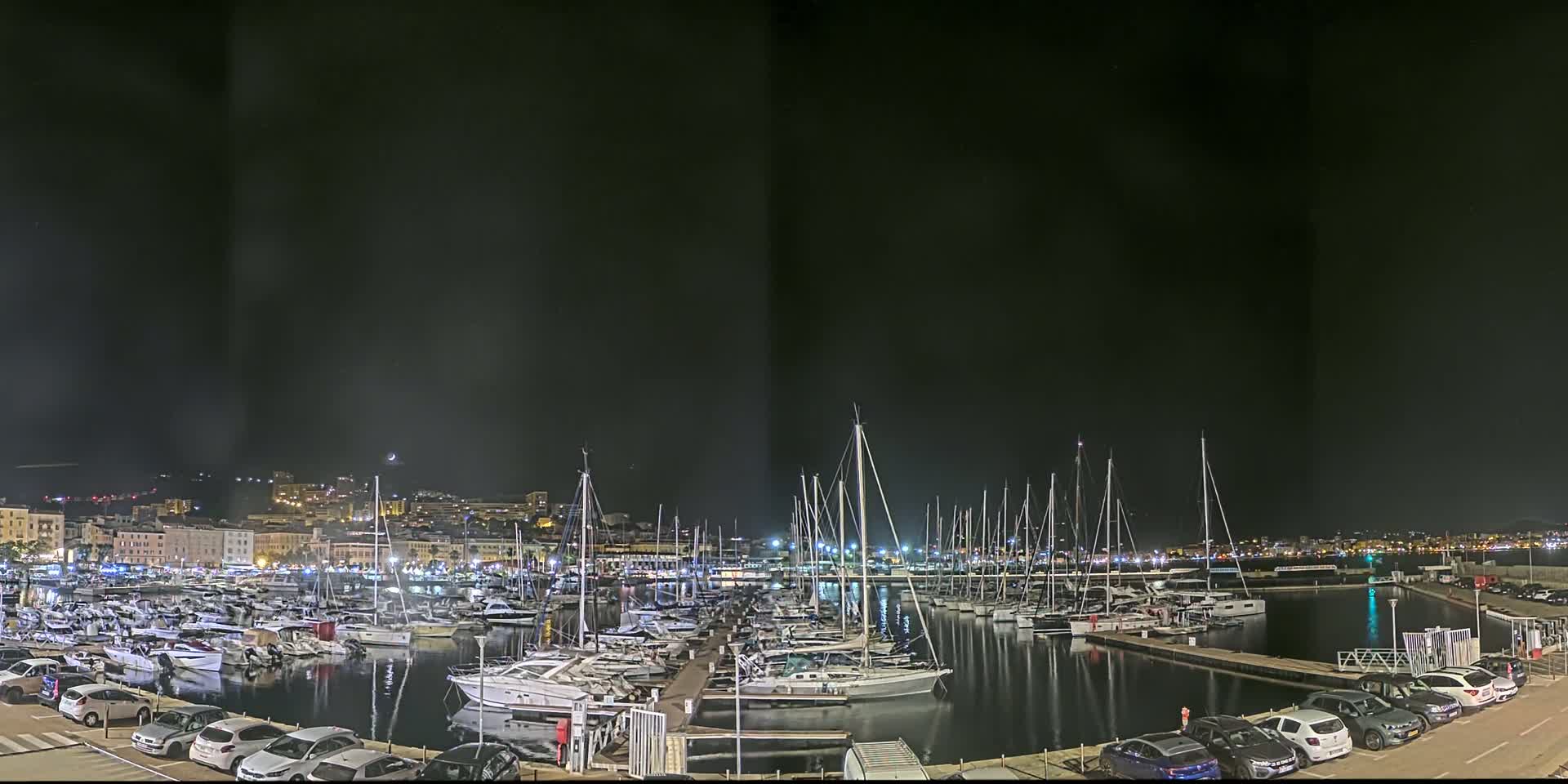 A nighttime view of a marina filled with sailboats and yachts, under a dark sky with a few lights illuminating the city in the background.