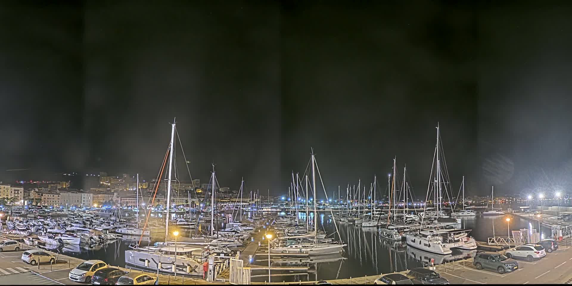 A nighttime view of a marina filled with sailboats and yachts, under a dark, slightly hazy sky, with cars parked in a lot nearby.