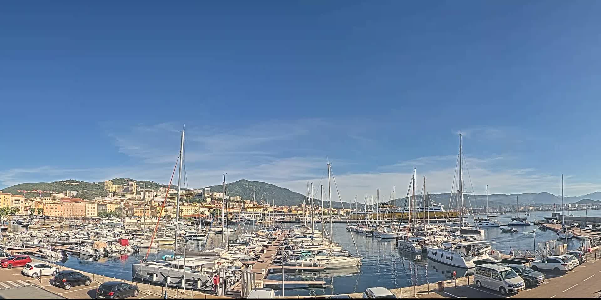A marina filled with sailboats and yachts is situated in front of a city nestled against a mountain range under a clear blue sky.