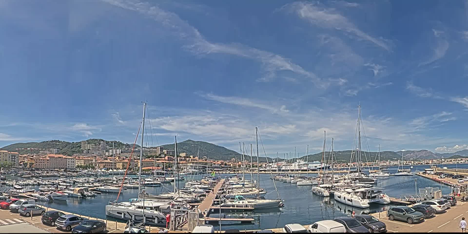 A marina filled with sailboats and yachts is situated in front of a town nestled against a mountain range under a mostly sunny sky with a few scattered clouds.
