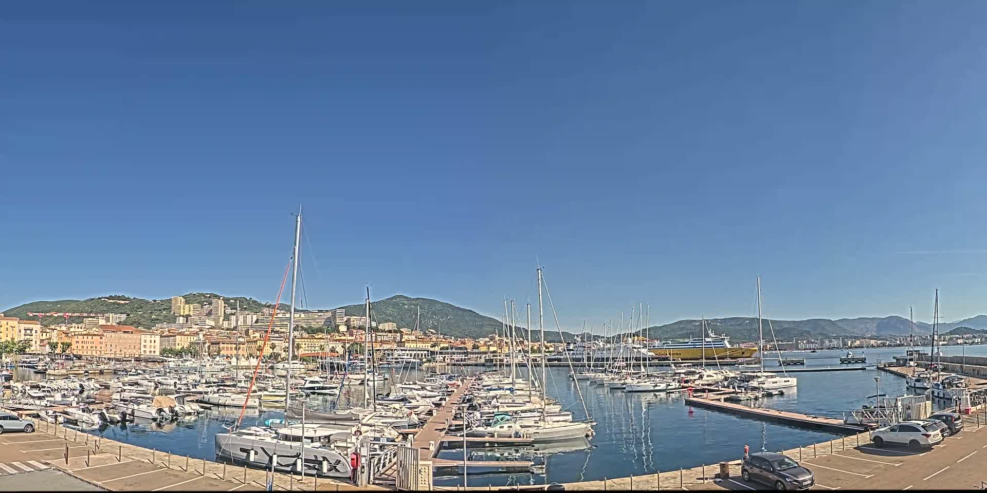 A marina filled with numerous sailboats and yachts is situated in front of a city nestled against a mountain range under a clear blue sky.