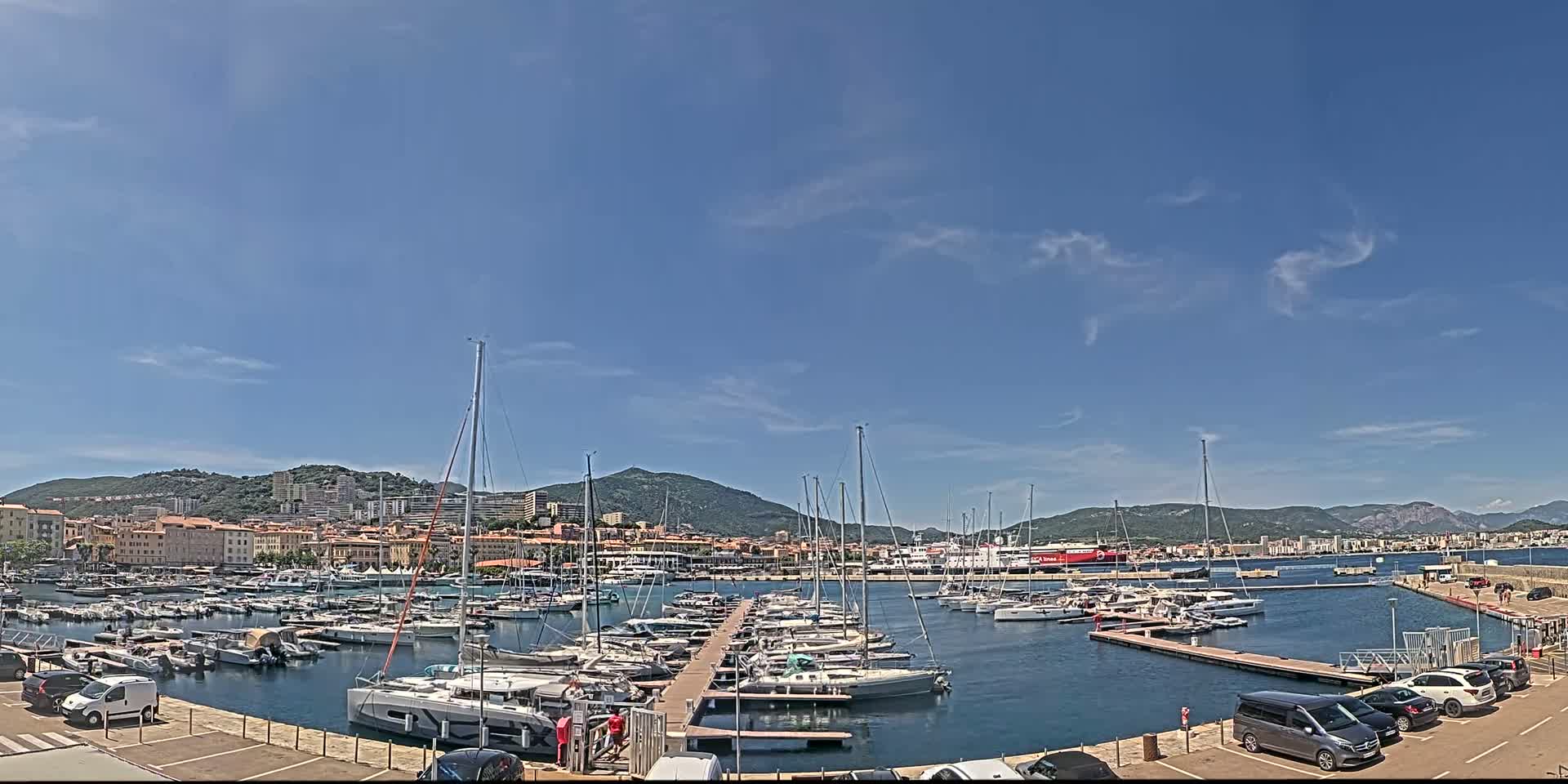 A marina filled with numerous sailboats and motorboats is situated in front of a town nestled against a mountain range under a mostly clear, sunny sky.