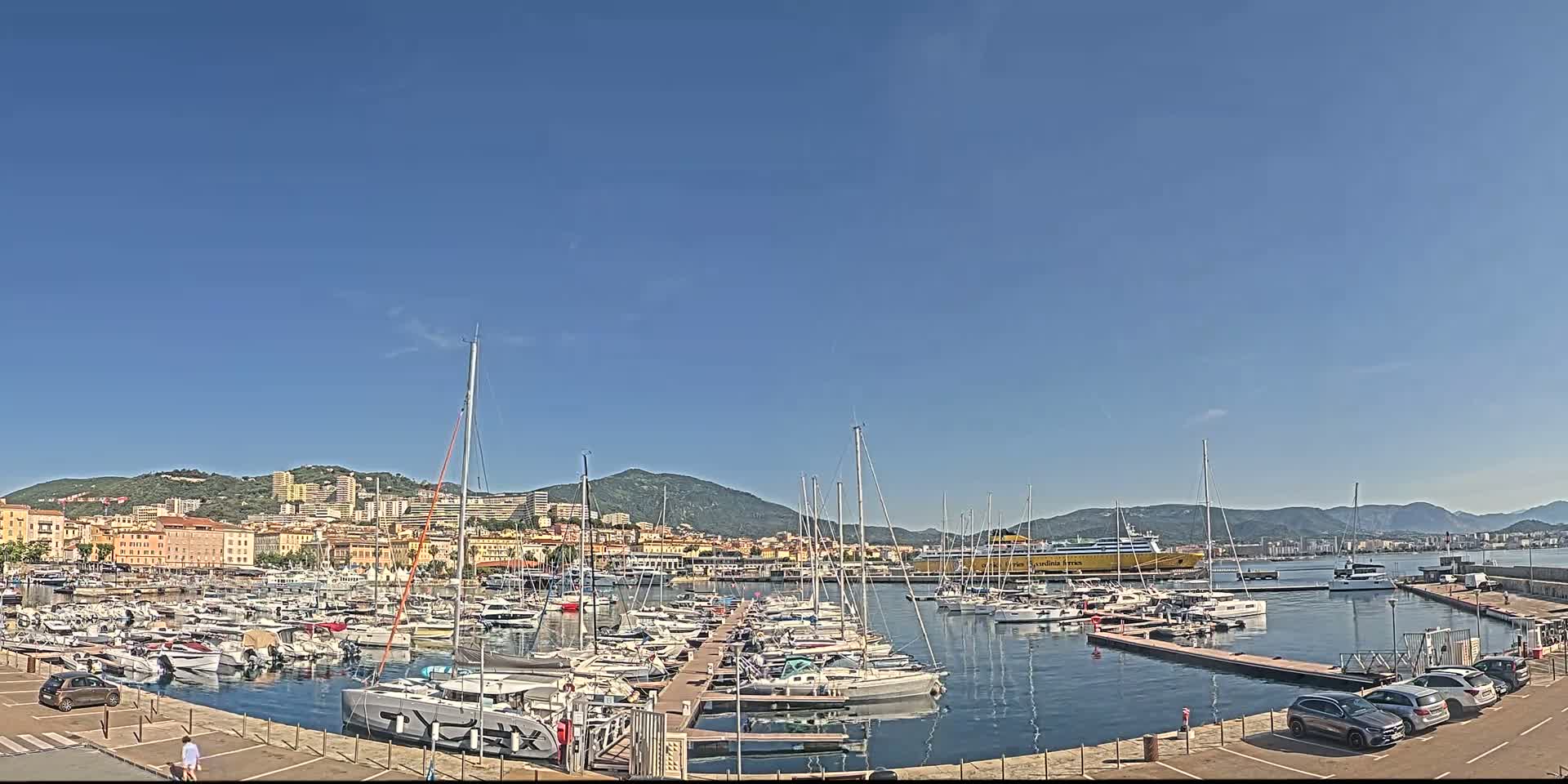 A marina filled with sailboats and yachts is situated in front of a city nestled against mountains under a clear blue sky.