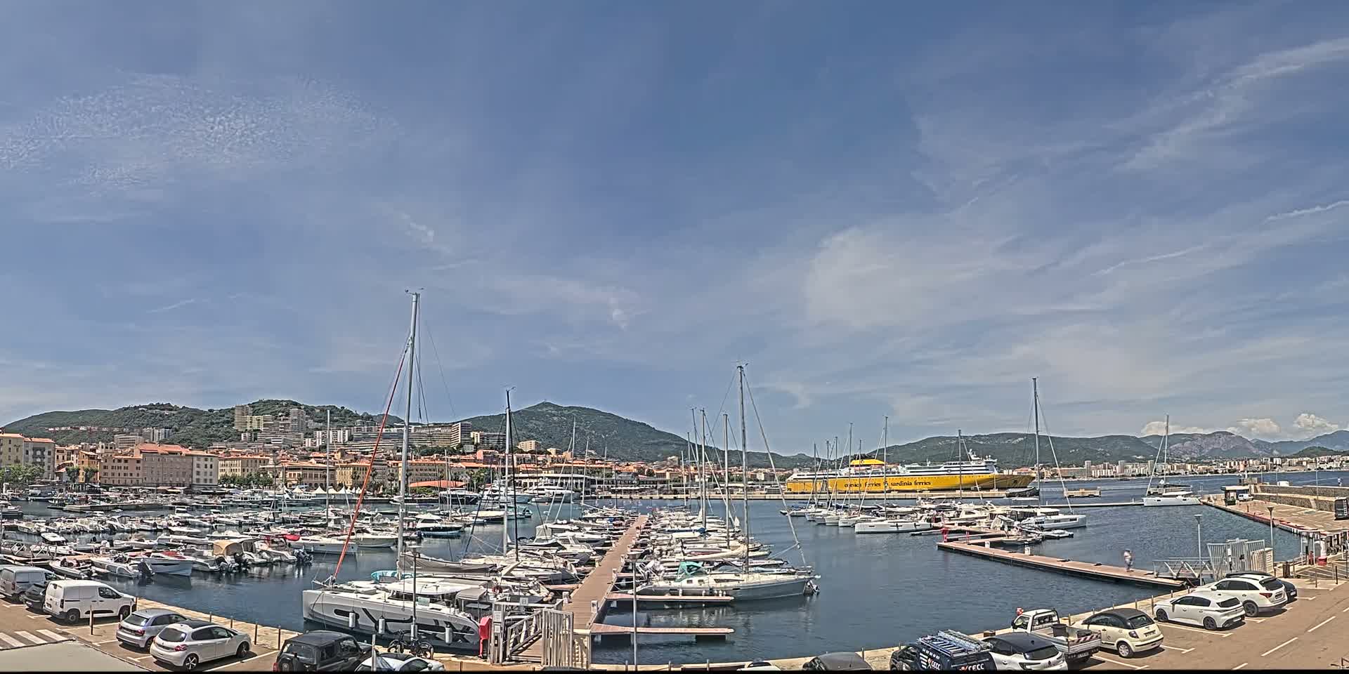 A marina filled with numerous sailboats and yachts is situated in front of a town nestled against a mountain range under a mostly clear, sunny sky.
