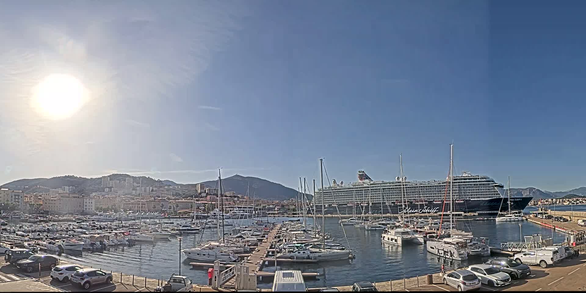 A large cruise ship is docked in a harbor filled with numerous smaller boats, under a bright, sunny sky and clear blue skies.
