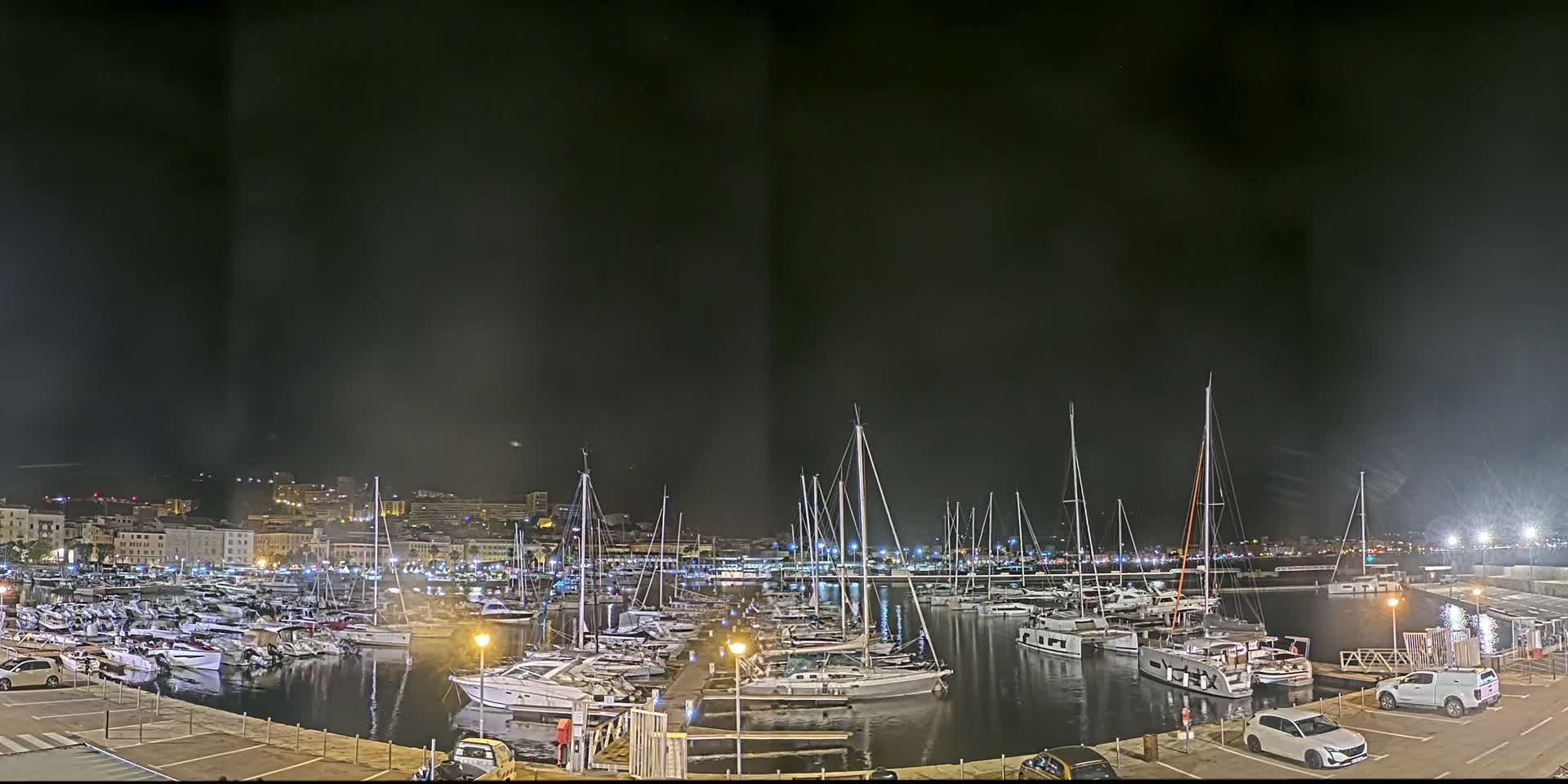 A marina filled with numerous boats is shown at night under a dark, cloudy sky, with a city skyline visible in the background.