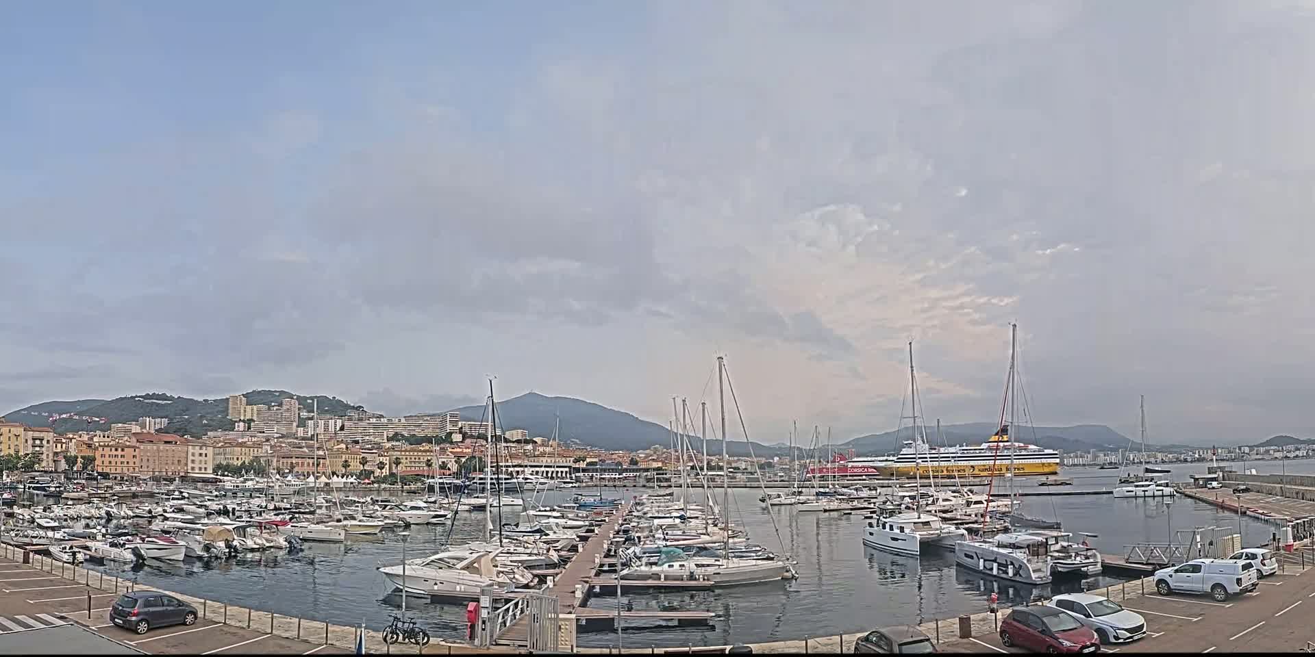 A marina filled with sailboats and motorboats is situated in front of a city nestled against mountains under a mostly cloudy sky.