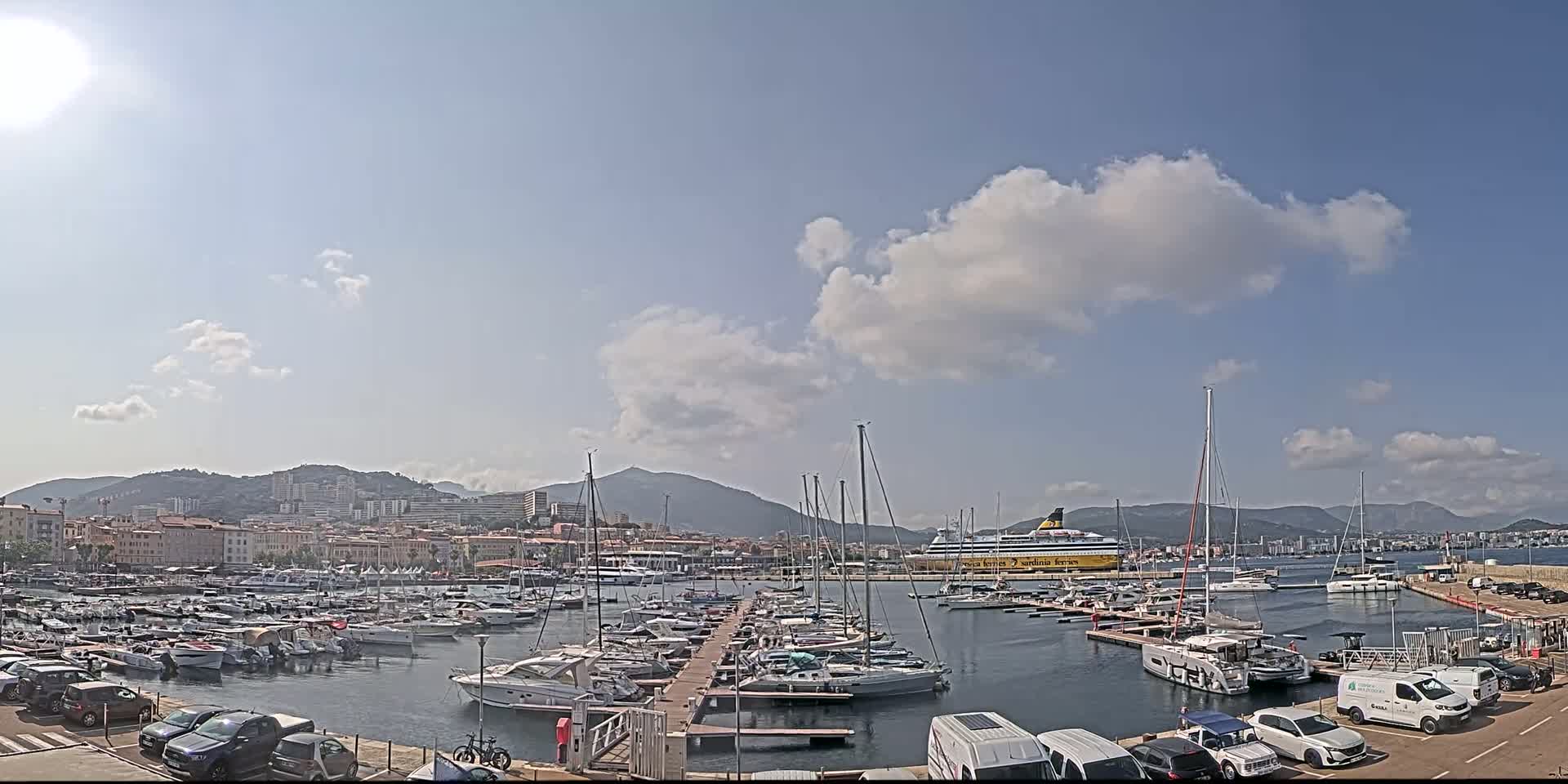 A sunny day reveals a harbor filled with numerous boats and yachts, docked alongside a larger ferry, with mountains and a cityscape in the background.
