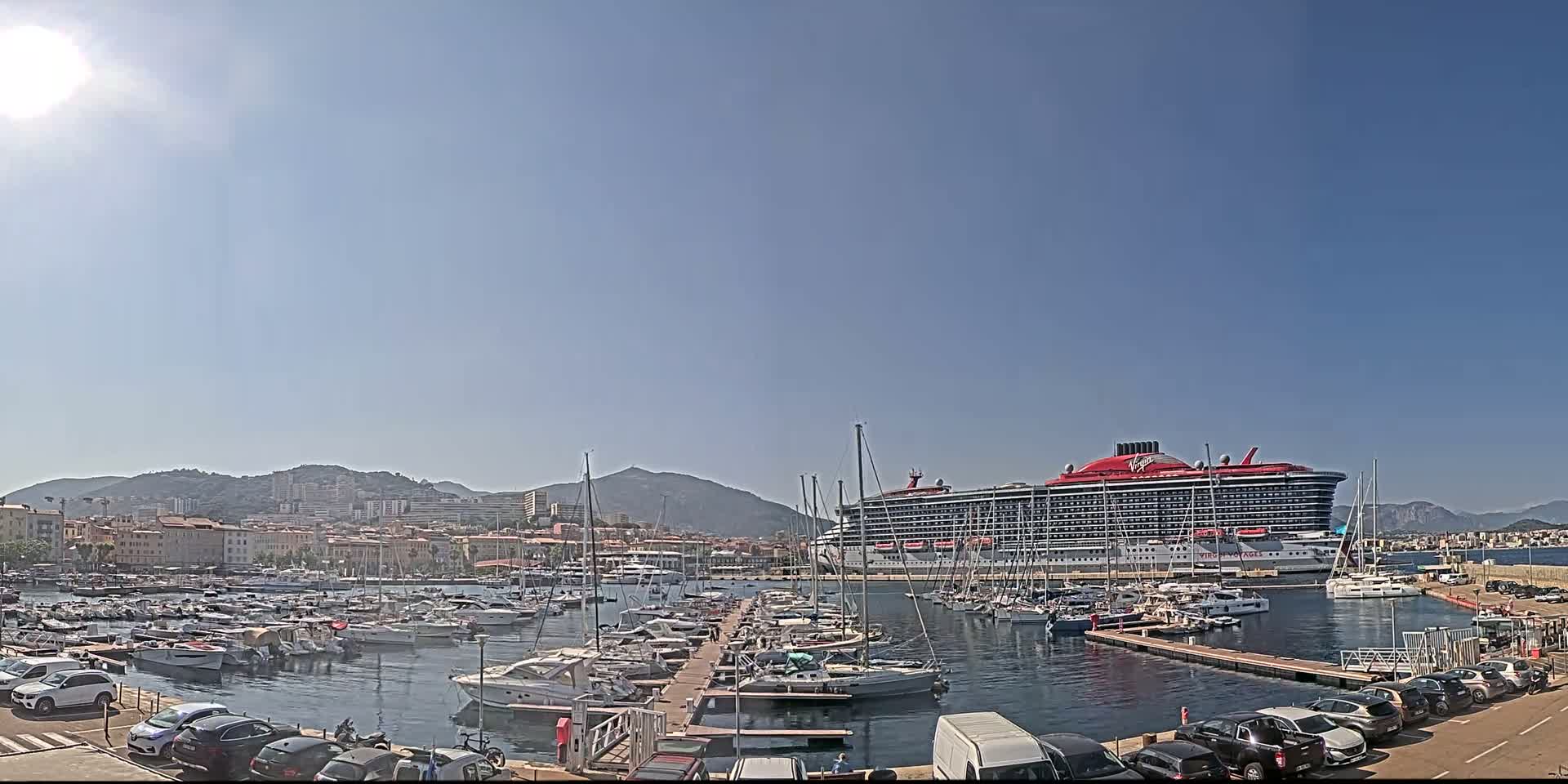 A large red cruise ship is docked in a harbor filled with many smaller boats, under a clear, sunny sky, with a town and mountains visible in the background.