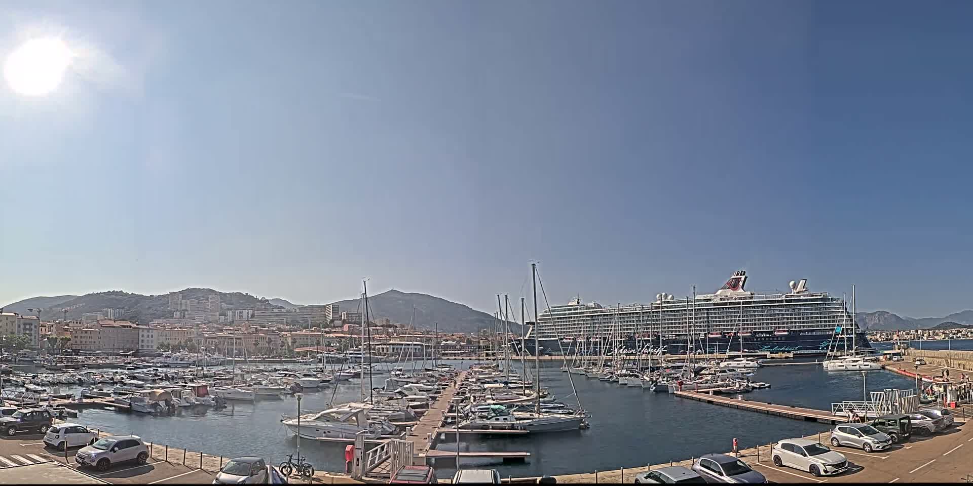 A large cruise ship is docked at a marina filled with smaller boats, under a bright, sunny sky; a city and mountains are visible in the background.