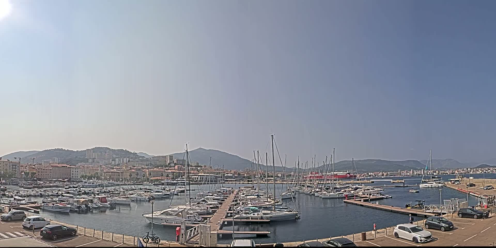 A sunny day reveals a harbor filled with numerous boats and yachts, backed by a cityscape nestled against mountains.