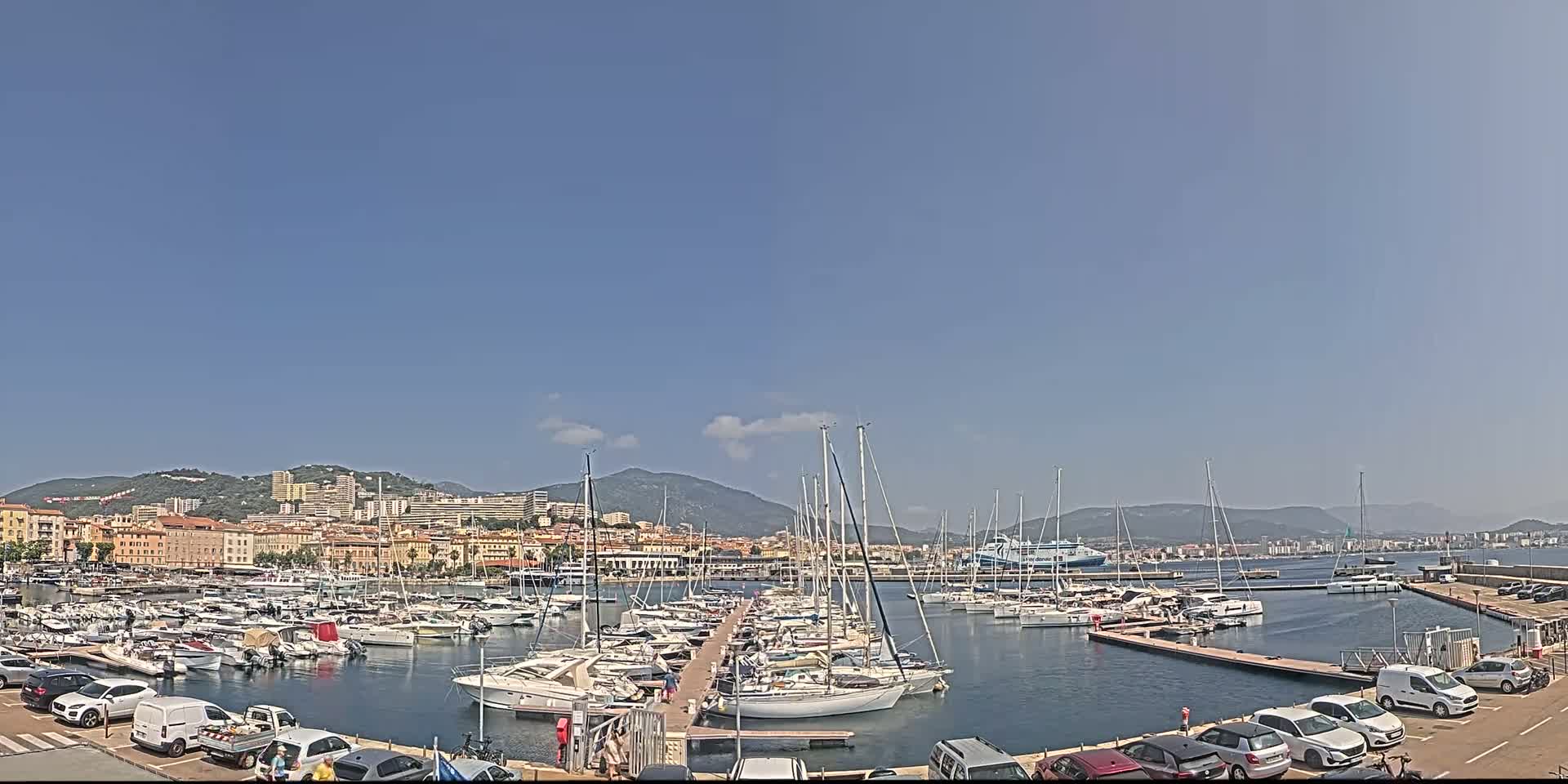 A marina filled with sailboats and motorboats is situated in front of a city nestled against mountains under a clear, sunny sky.