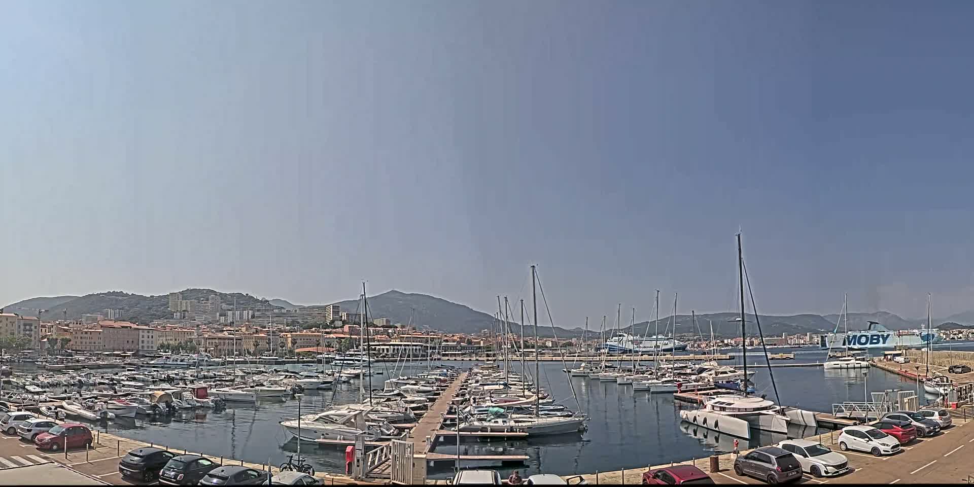 A marina filled with numerous sailboats and yachts is situated near a city under a clear, sunny sky, with mountains visible in the background.