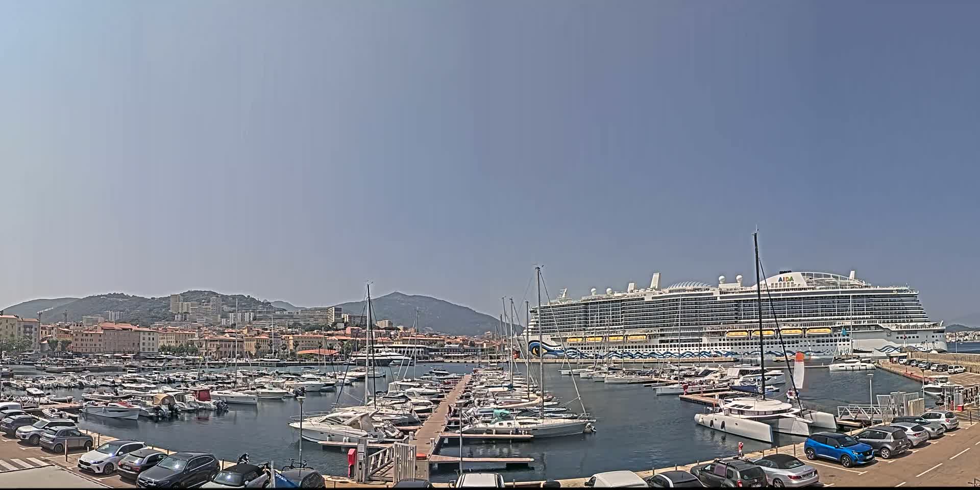 A large cruise ship is docked in a harbor filled with numerous yachts and boats, under a clear, sunny sky with mountains in the background.