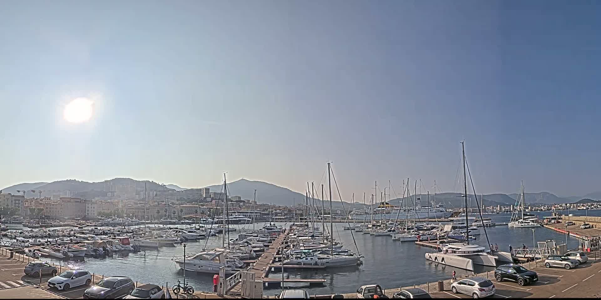 A sunny day shows a harbor filled with many sailboats and yachts, docked near a city with mountains in the background.