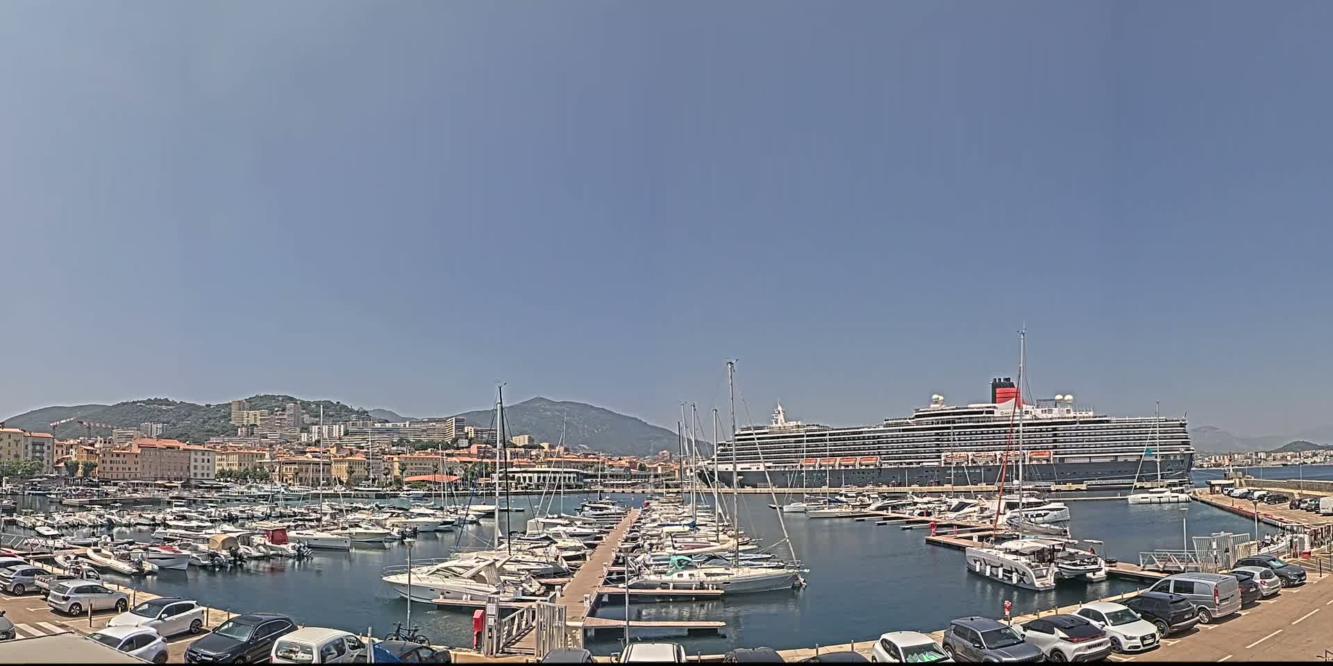 A large cruise ship is docked in a harbor filled with numerous smaller boats, under a clear, sunny sky.
