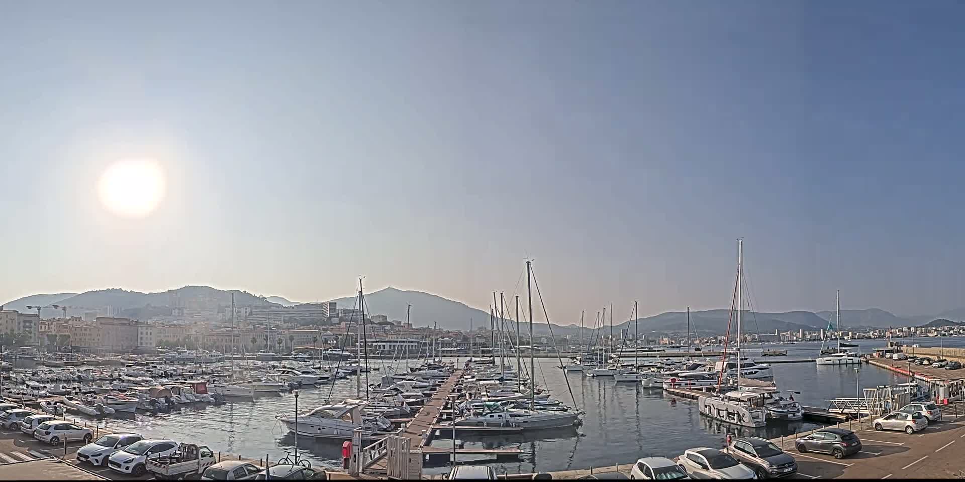 A sunny day reveals a harbor filled with numerous boats and yachts docked at piers, with a city and mountains in the background.