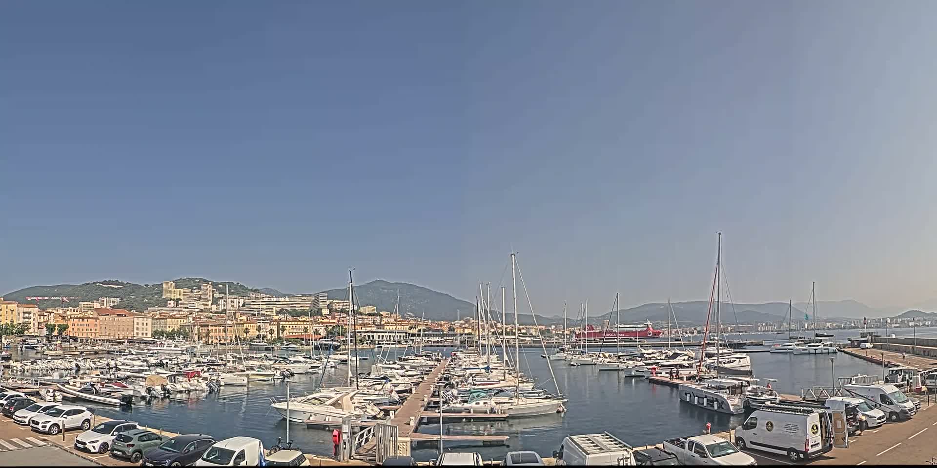 A harbor filled with numerous sailboats and yachts is situated in front of a city nestled against a mountain range under a clear, sunny sky.