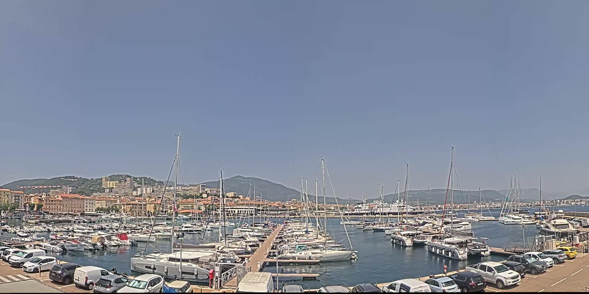 A marina filled with sailboats and motorboats is situated in front of a city nestled against a mountain range under a clear, bright sky.