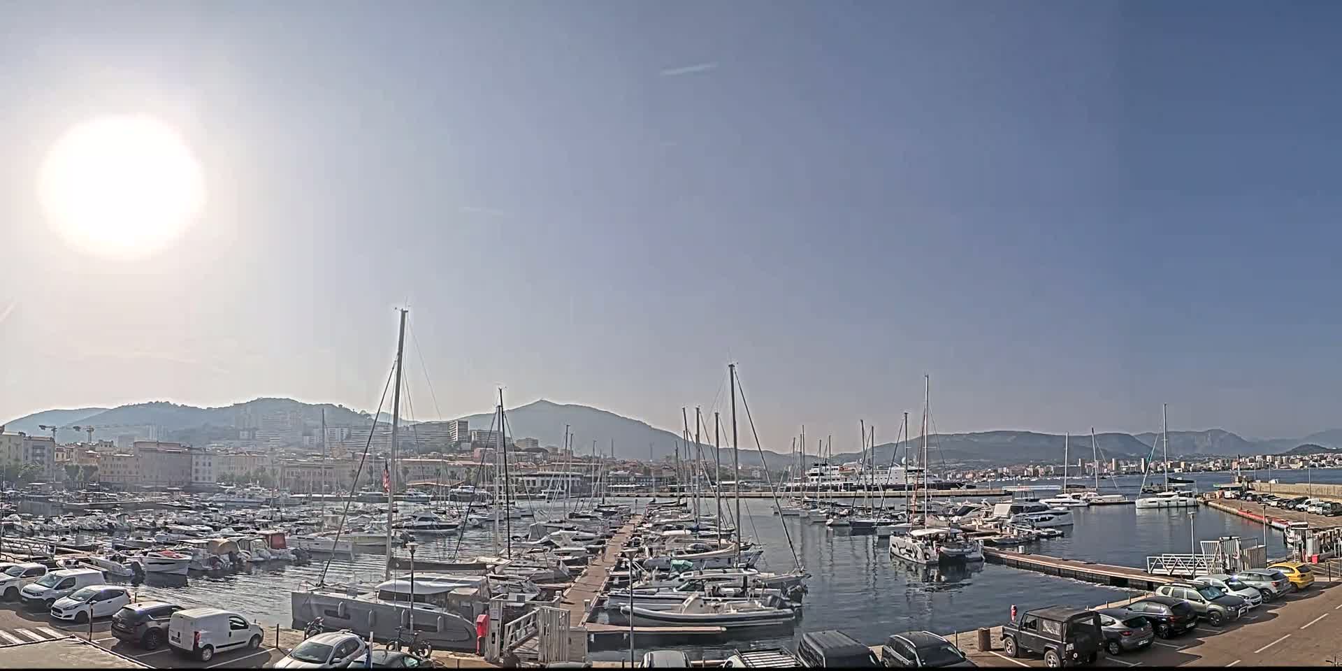 A sunny day at a marina filled with sailboats and yachts, backed by a city and mountains.