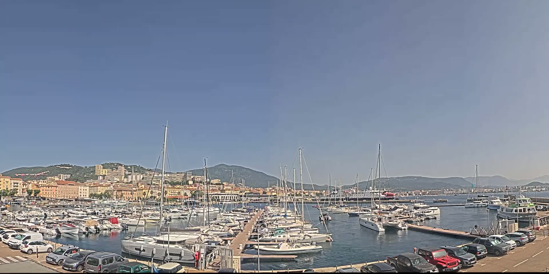A marina filled with numerous sailboats and motorboats is nestled in a harbor, with a city and hills in the background under a clear blue sky.