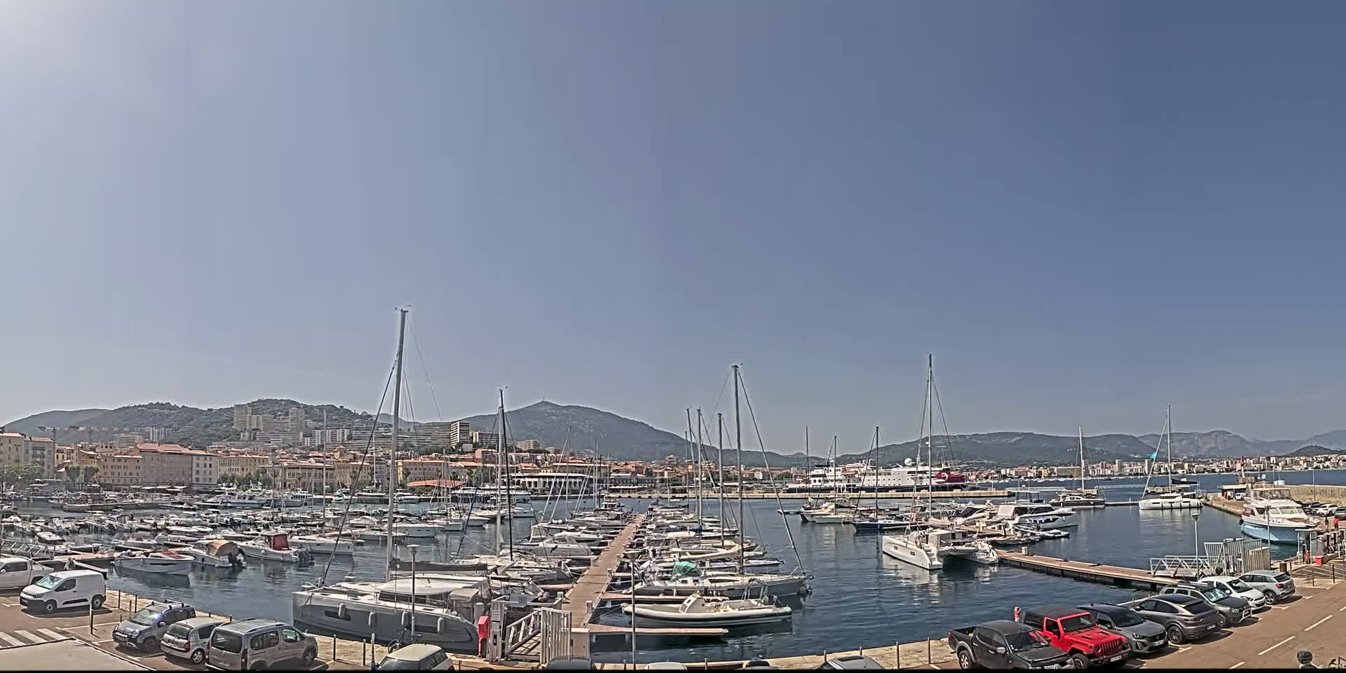 A marina filled with numerous sailboats and motorboats is situated in front of a town nestled against a mountain range under a clear, sunny sky.