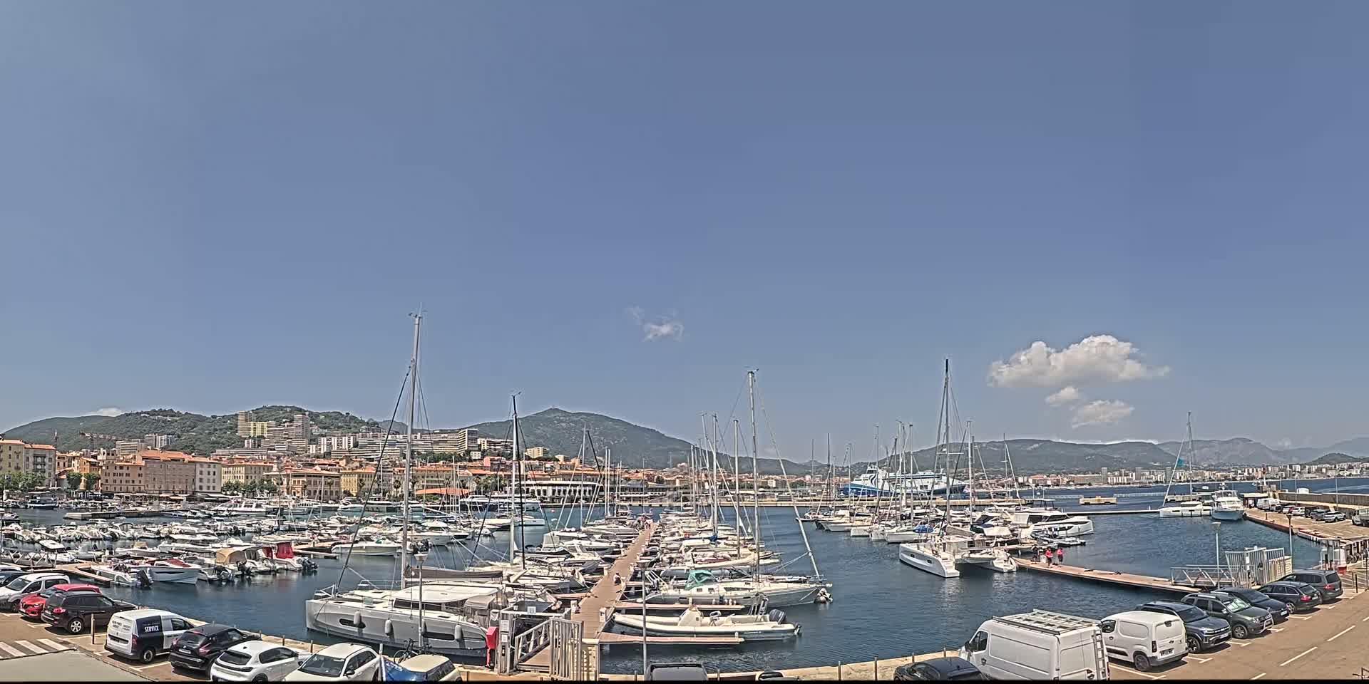 A marina filled with numerous sailboats and motorboats is situated near a town backed by mountains under a mostly clear, sunny sky.