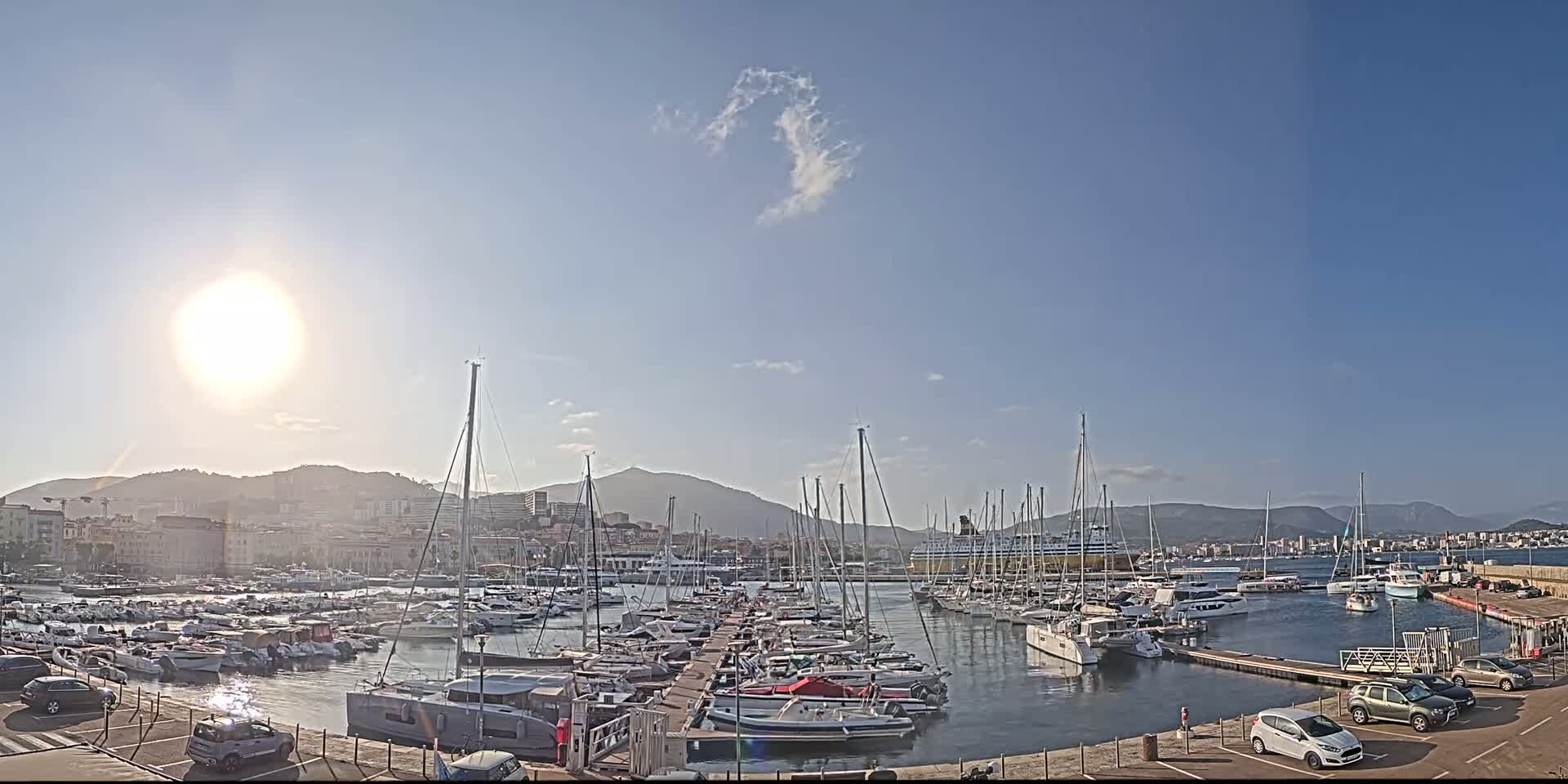 A sunny day shows a harbor filled with numerous sailboats and yachts, docked along a pier with cars parked nearby, mountains in the background.