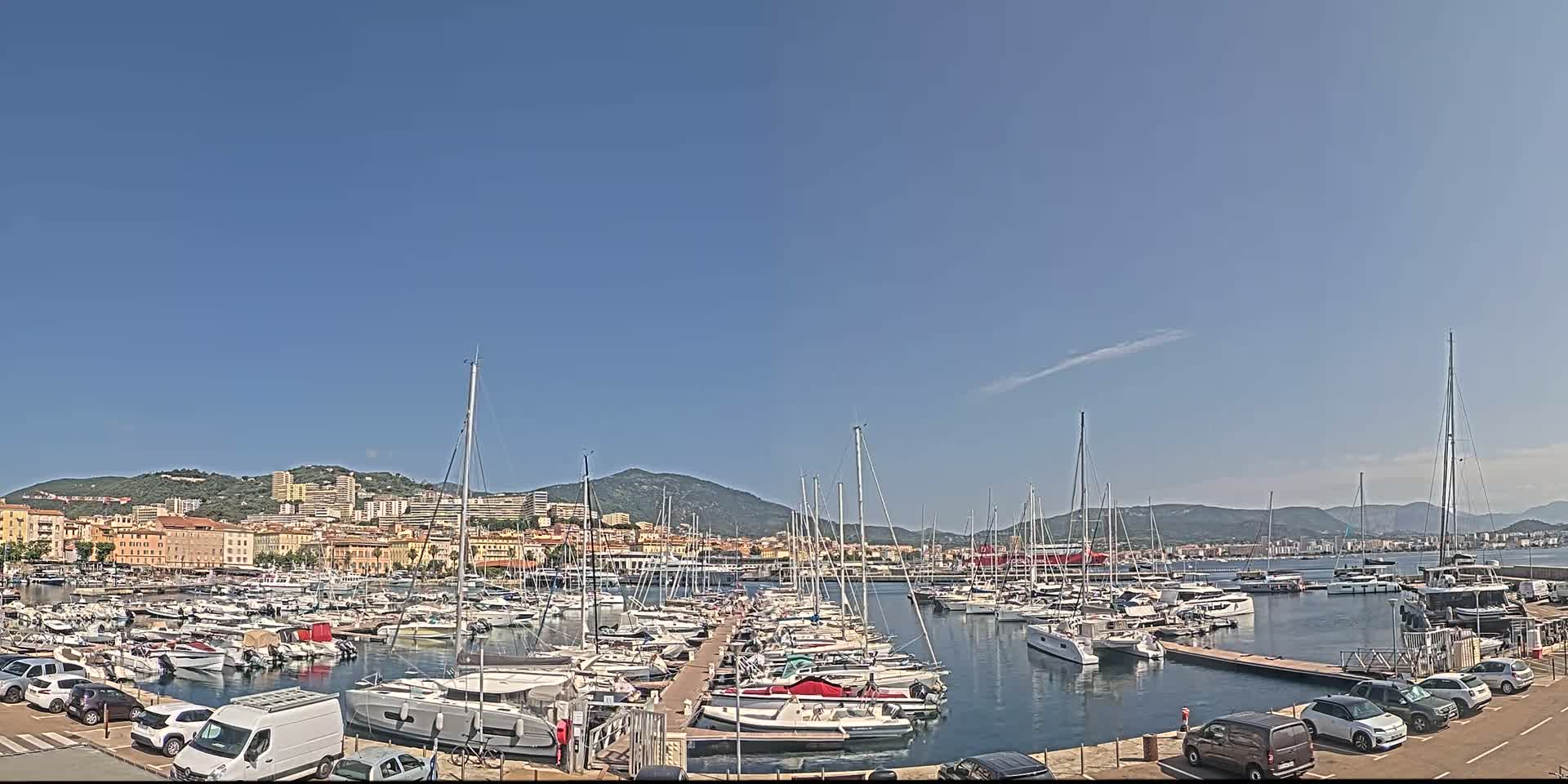 A marina filled with numerous sailboats and motorboats is situated near a town nestled against a mountain range under a clear blue sky.