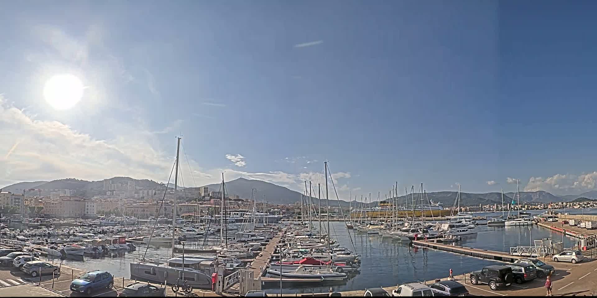 A sunny day reveals a marina filled with numerous sailboats and yachts, backed by a city and mountains.