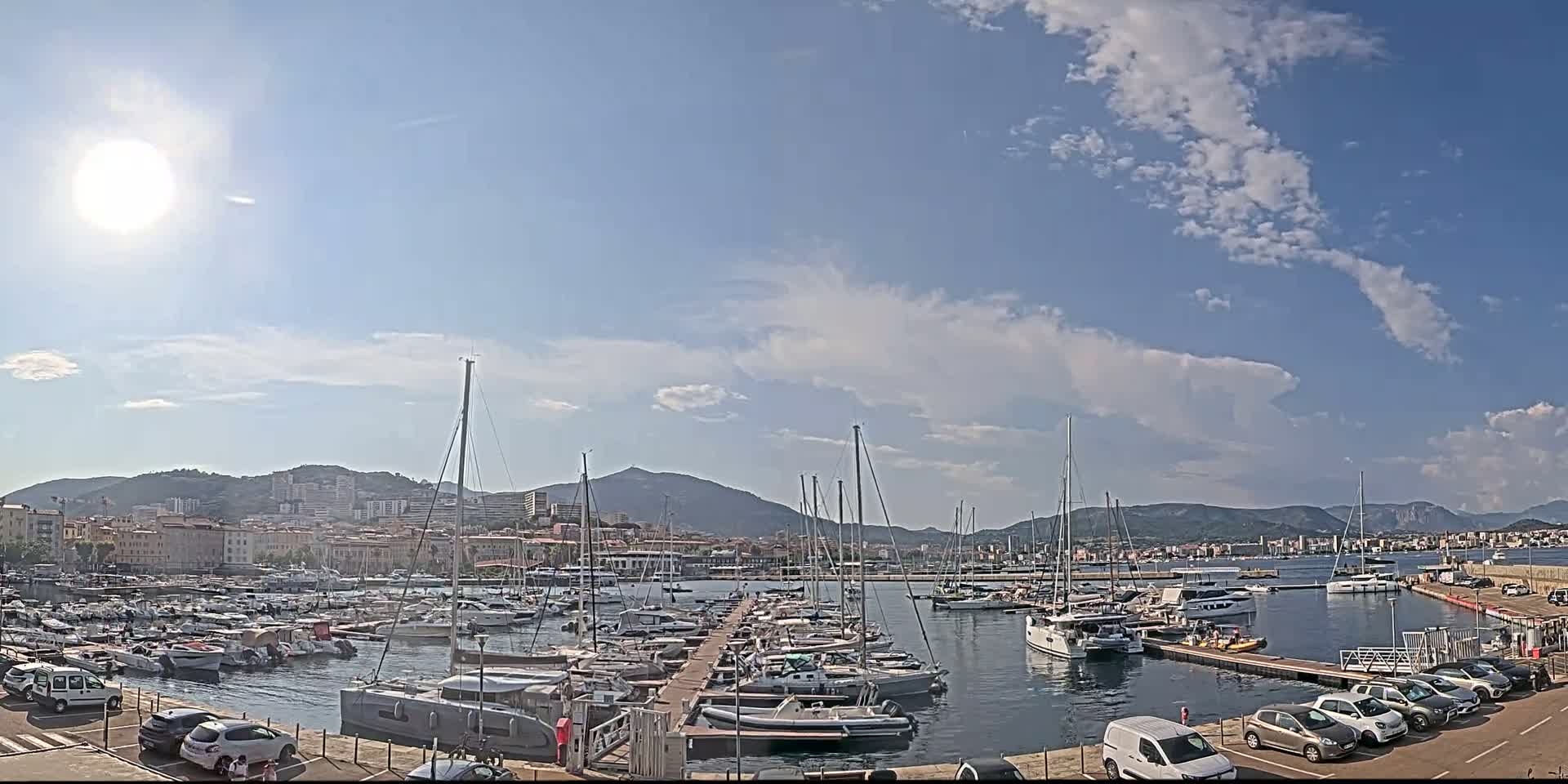 A sunny day with partly cloudy skies over a marina filled with sailboats and motorboats, backed by a city and mountains.