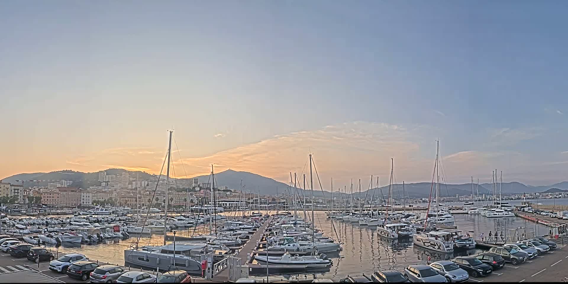 A marina filled with numerous sailboats and motorboats is situated near a city at sunset under a mostly clear sky.