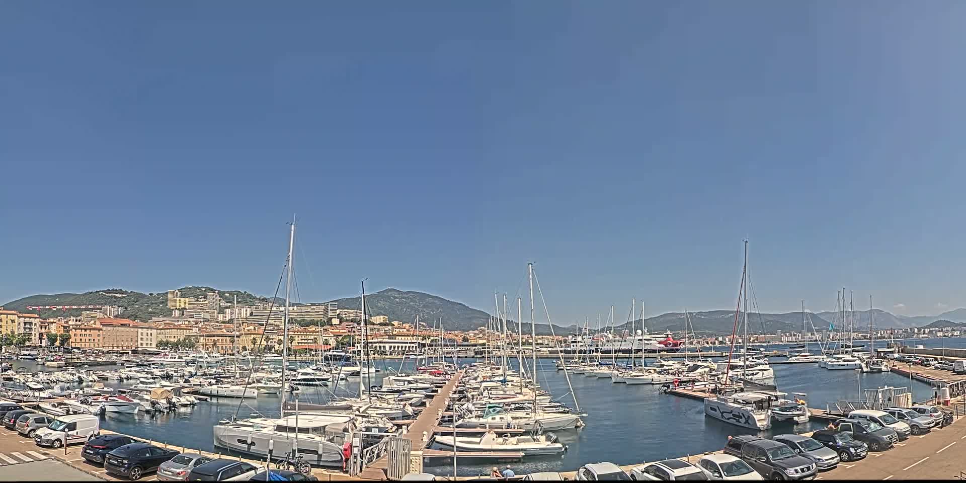 A marina filled with numerous sailboats and motorboats is situated near a city under a clear blue sky.