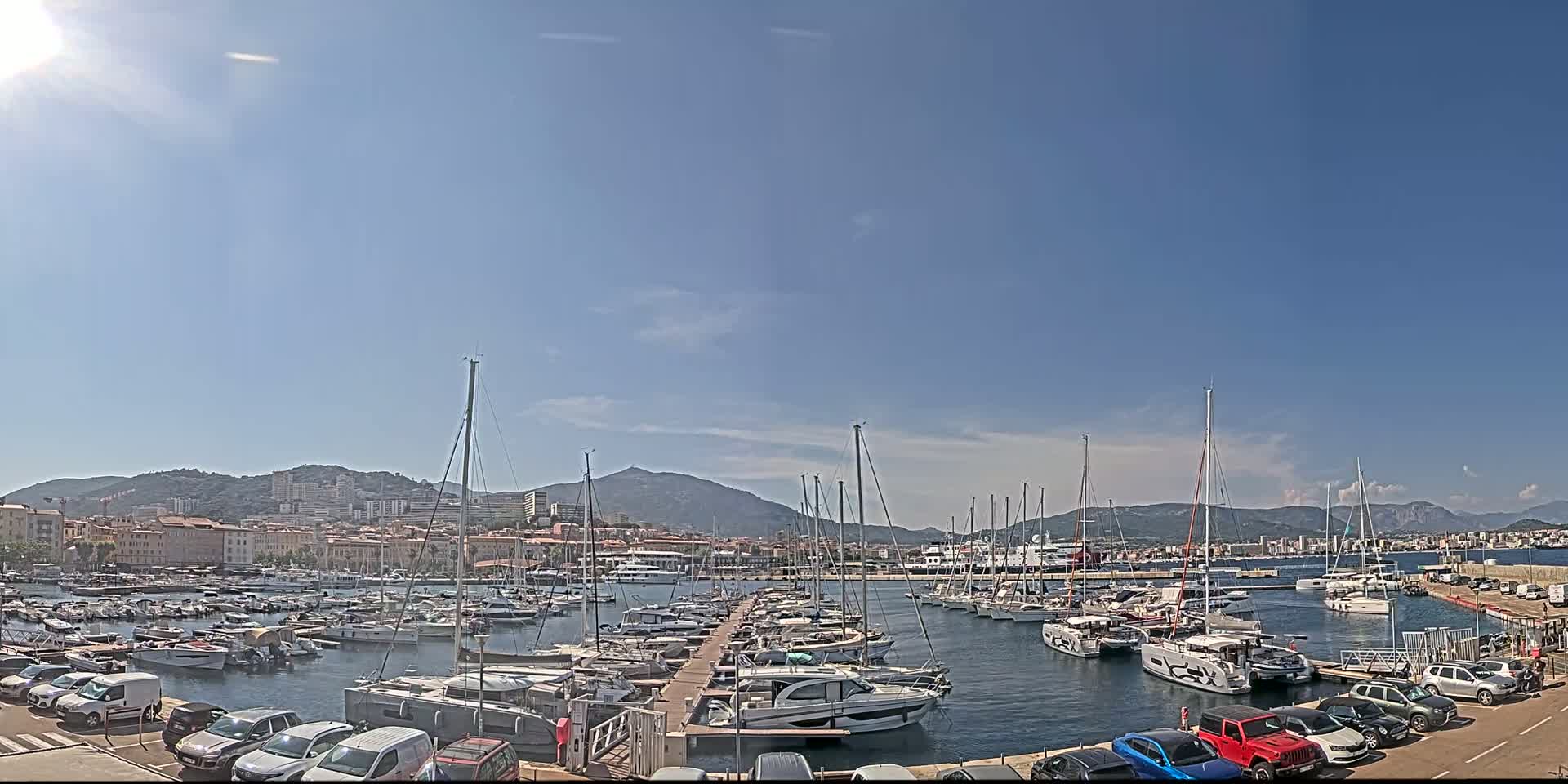 A sunny day shows a marina filled with sailboats and motorboats, docked next to a city backed by mountains.