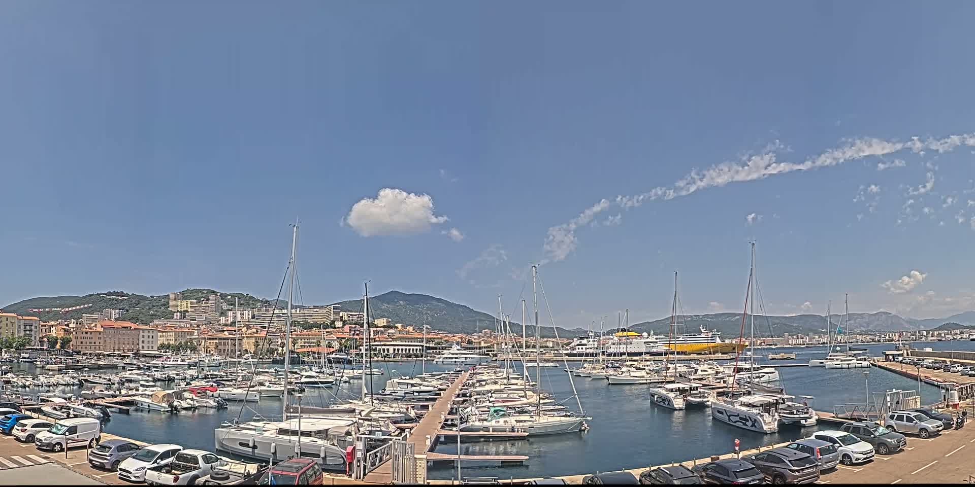 A marina filled with sailboats and motorboats is nestled in a harbor town, backed by hills under a mostly clear, sunny sky with a few scattered clouds.