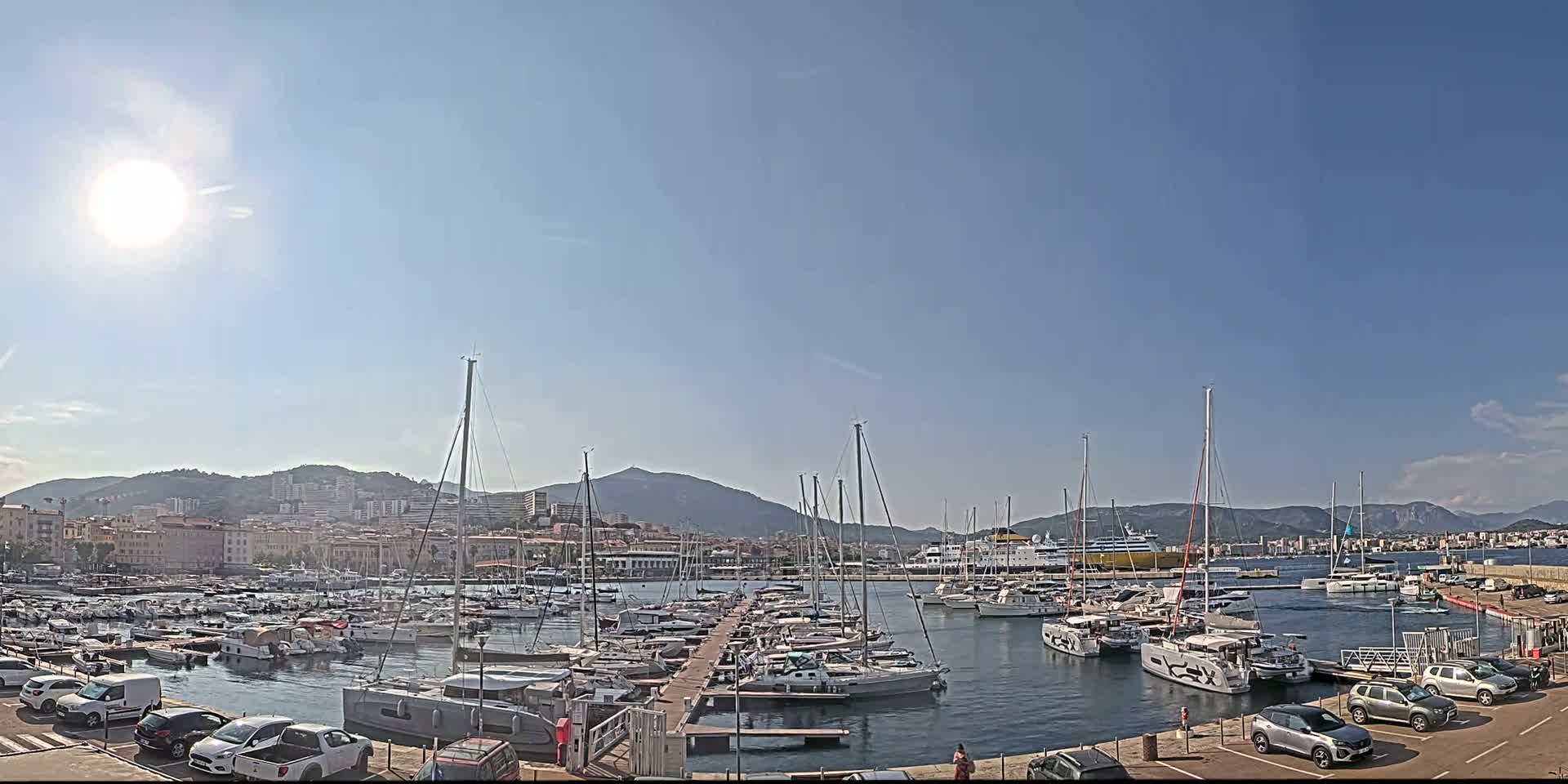 A sunny day shows a harbor filled with sailboats and yachts, a ferry in the distance, and cars parked along the shore with mountains in the background.