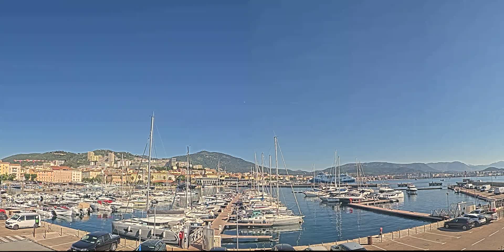 A marina filled with numerous sailboats and motorboats is situated in front of a city nestled against a mountain range under a clear blue sky.
