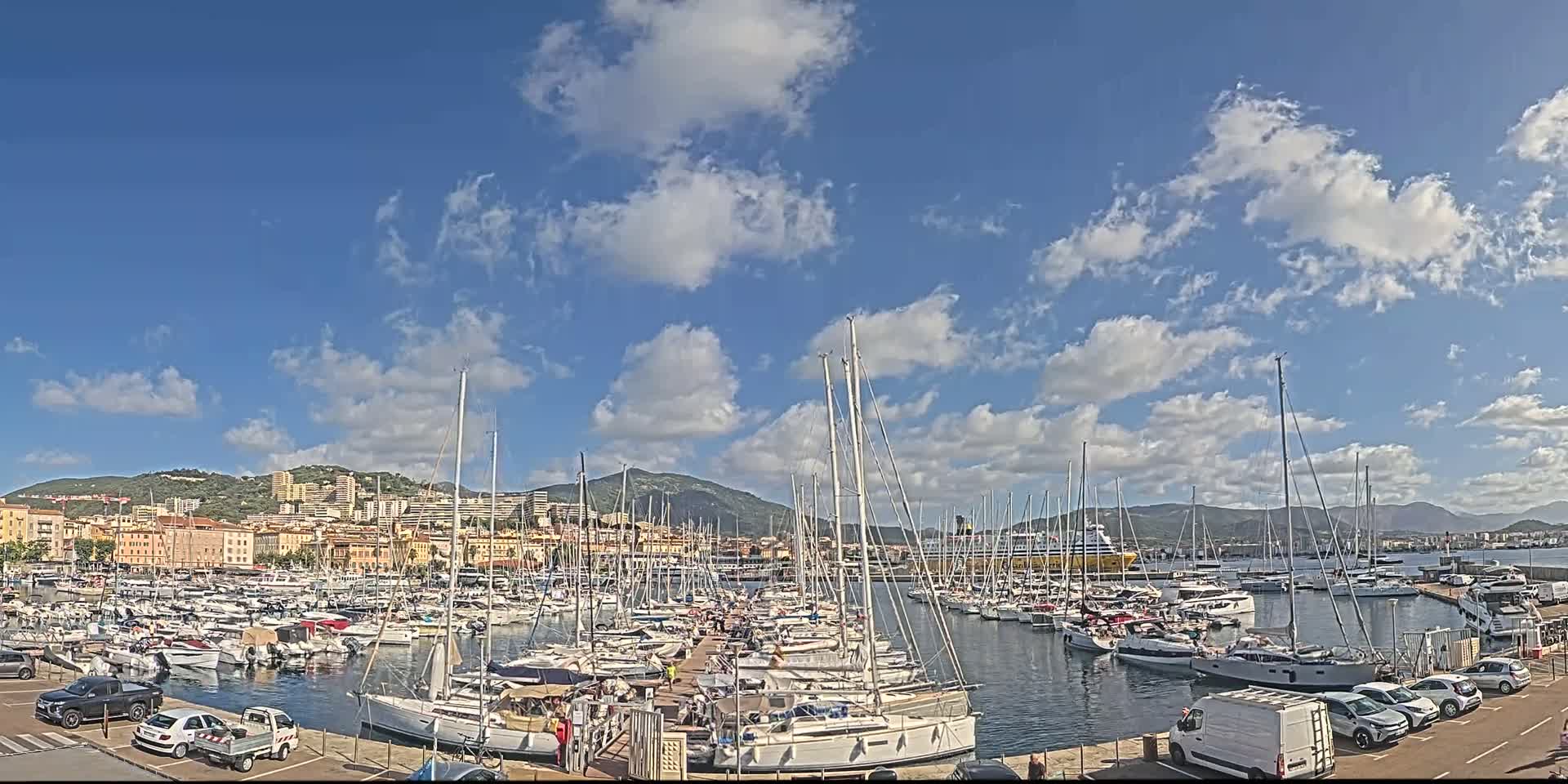 A marina filled with numerous sailboats and motorboats is situated in front of a city and hills under a mostly sunny sky with scattered cumulus clouds.