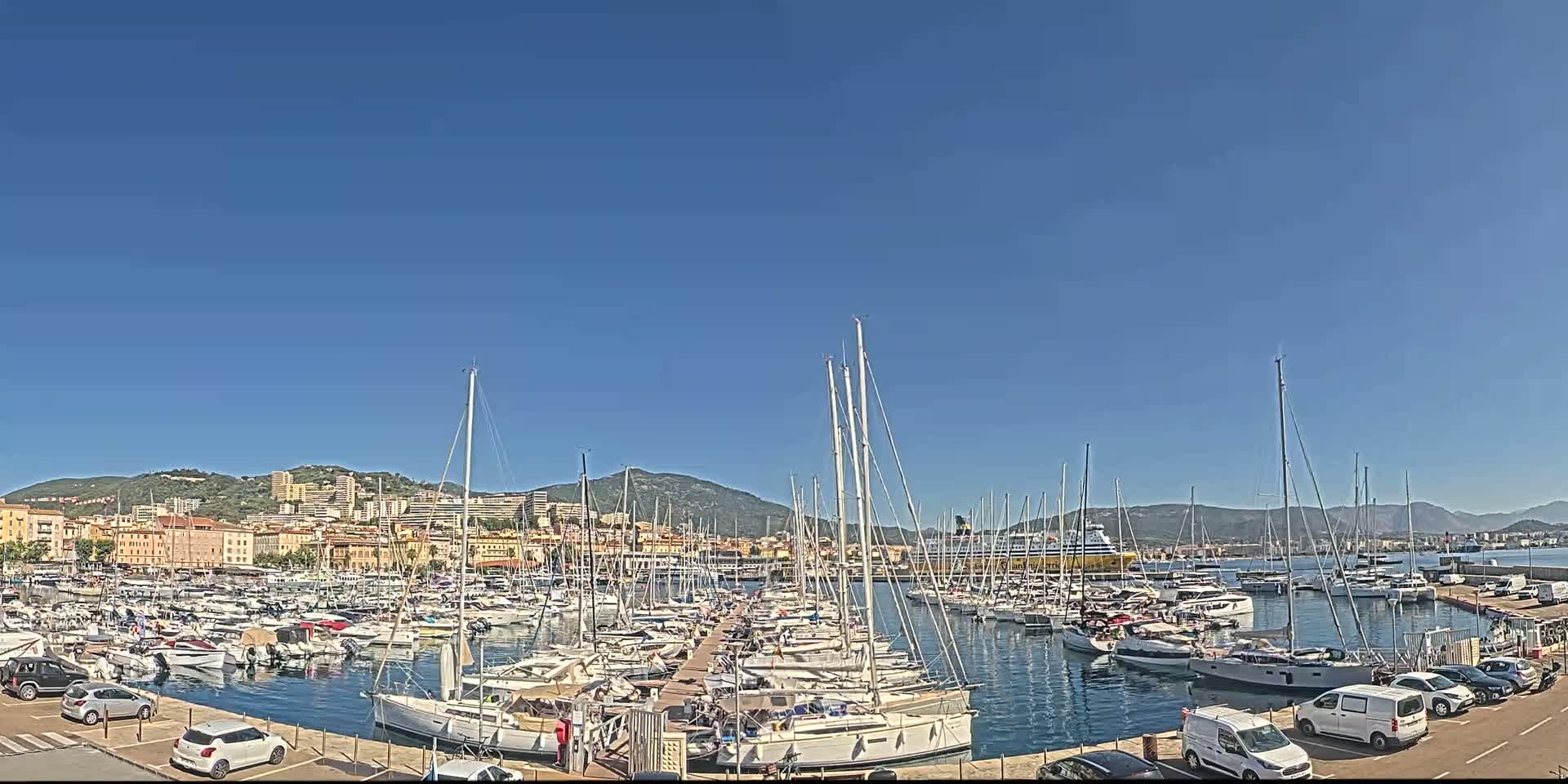 A marina filled with numerous sailboats and yachts is situated in front of a city nestled against a mountain range under a clear blue sky.