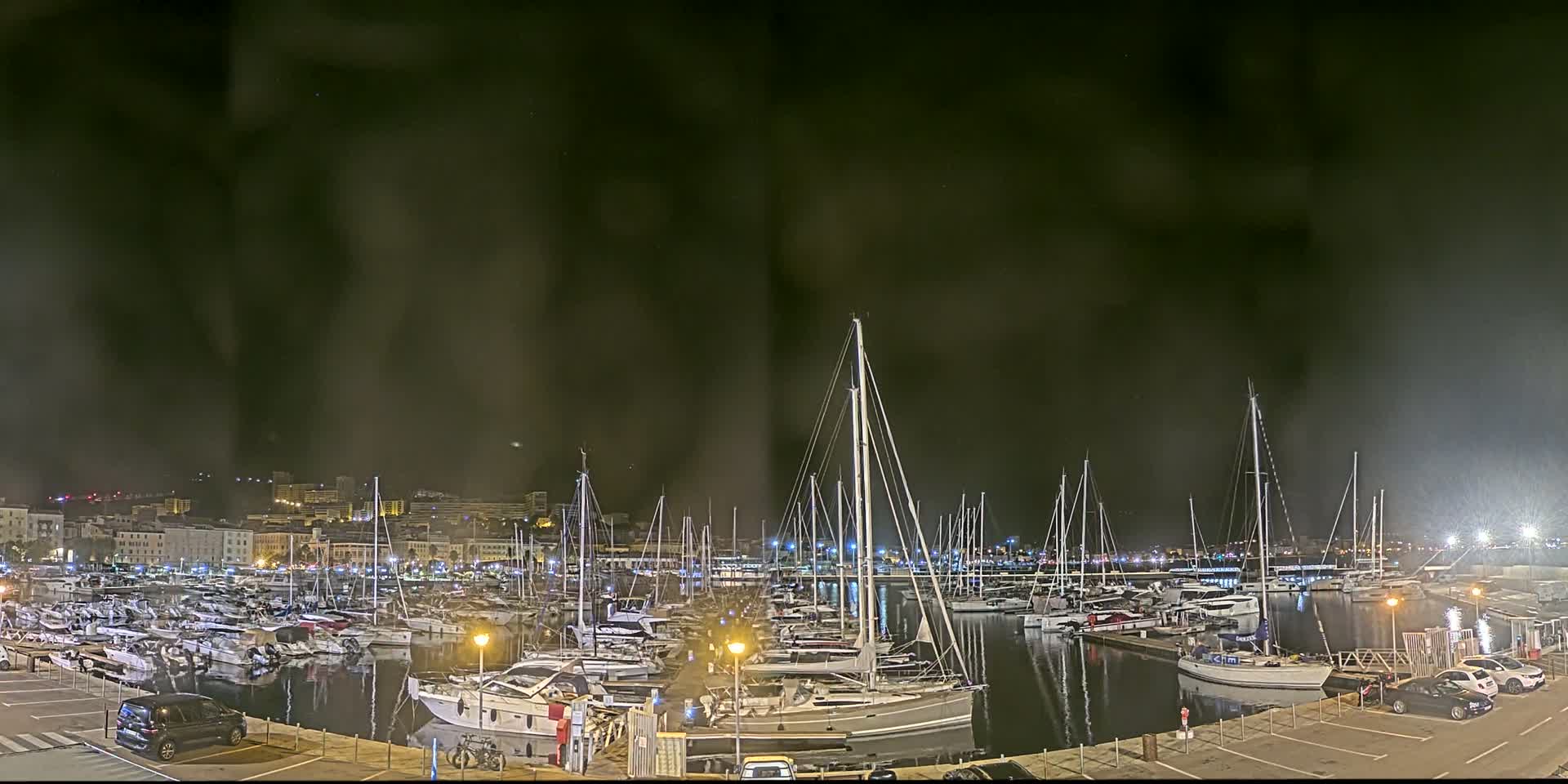 A marina filled with numerous sailboats and yachts is situated in front of a city nestled against a mountain range under a clear blue sky.