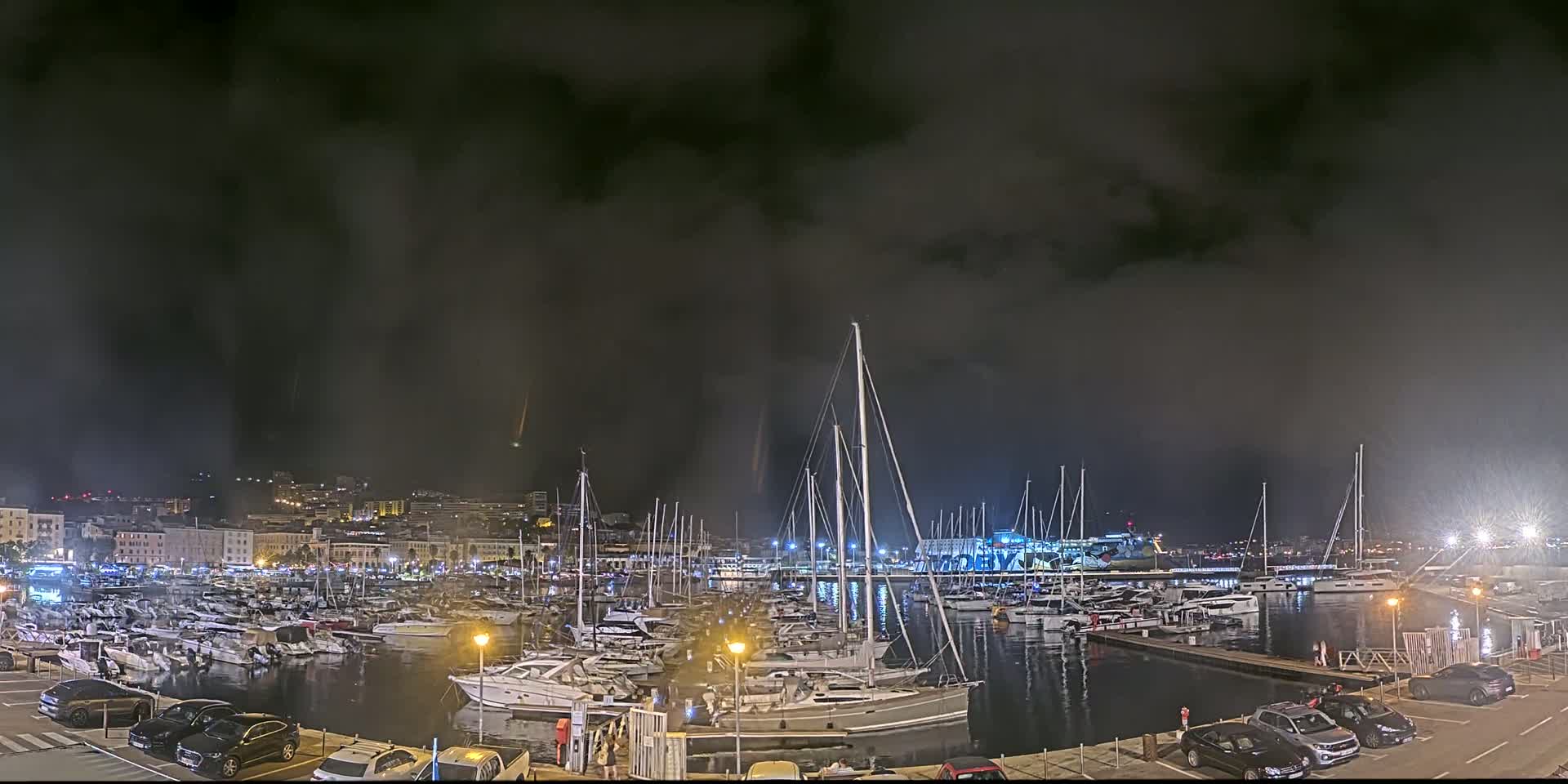 A nighttime view of a harbor filled with numerous boats and yachts under a dark, cloudy sky.