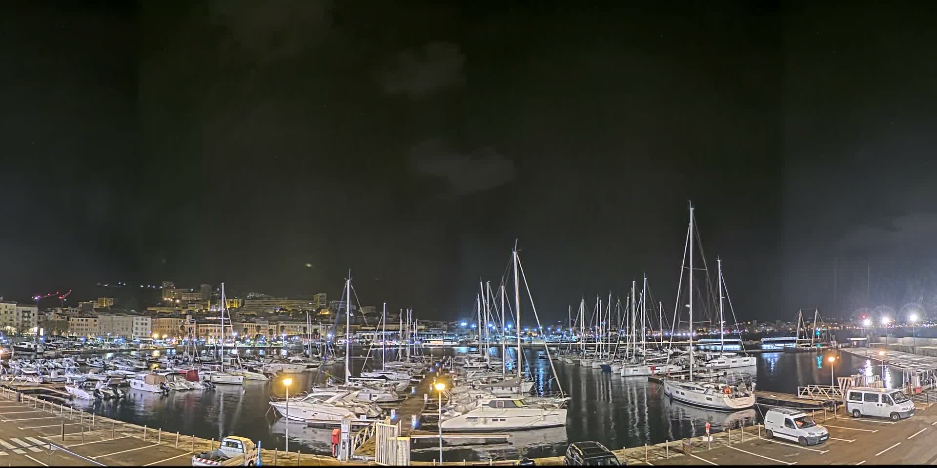 A wide view of a vibrant harbor at night features numerous sailboats and motorboats docked in calm waters, with a brightly lit city skyline in the background under an overcast sky.