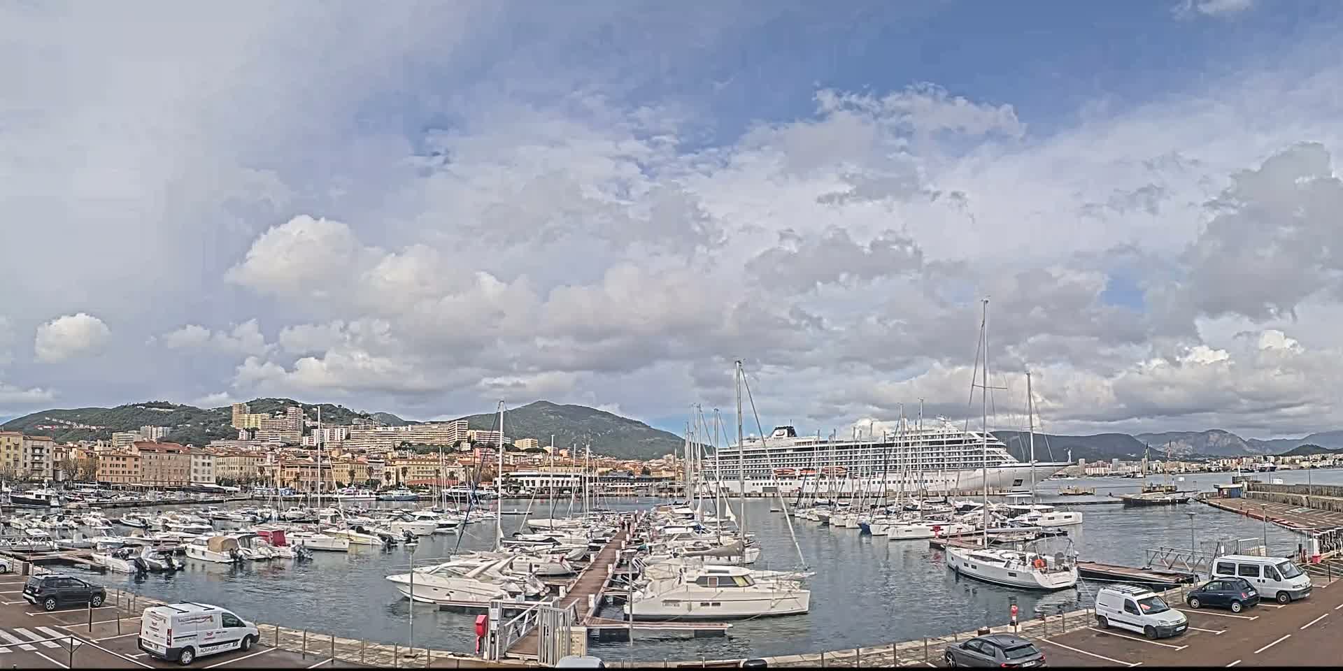 A bustling harbor filled with numerous yachts and sailboats is seen under a partly cloudy sky, with a large cruise ship docked in the background and a city nestled against green hills and distant mountains.