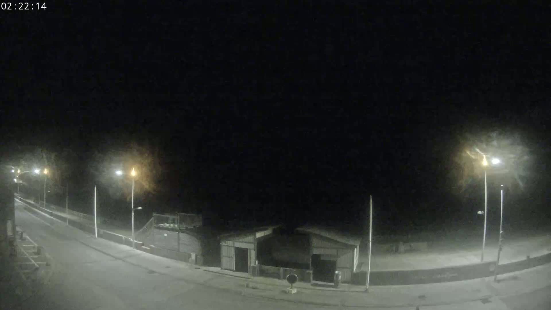 A nighttime, snowy scene shows a mostly empty road curving to the left, past some small buildings and streetlights.