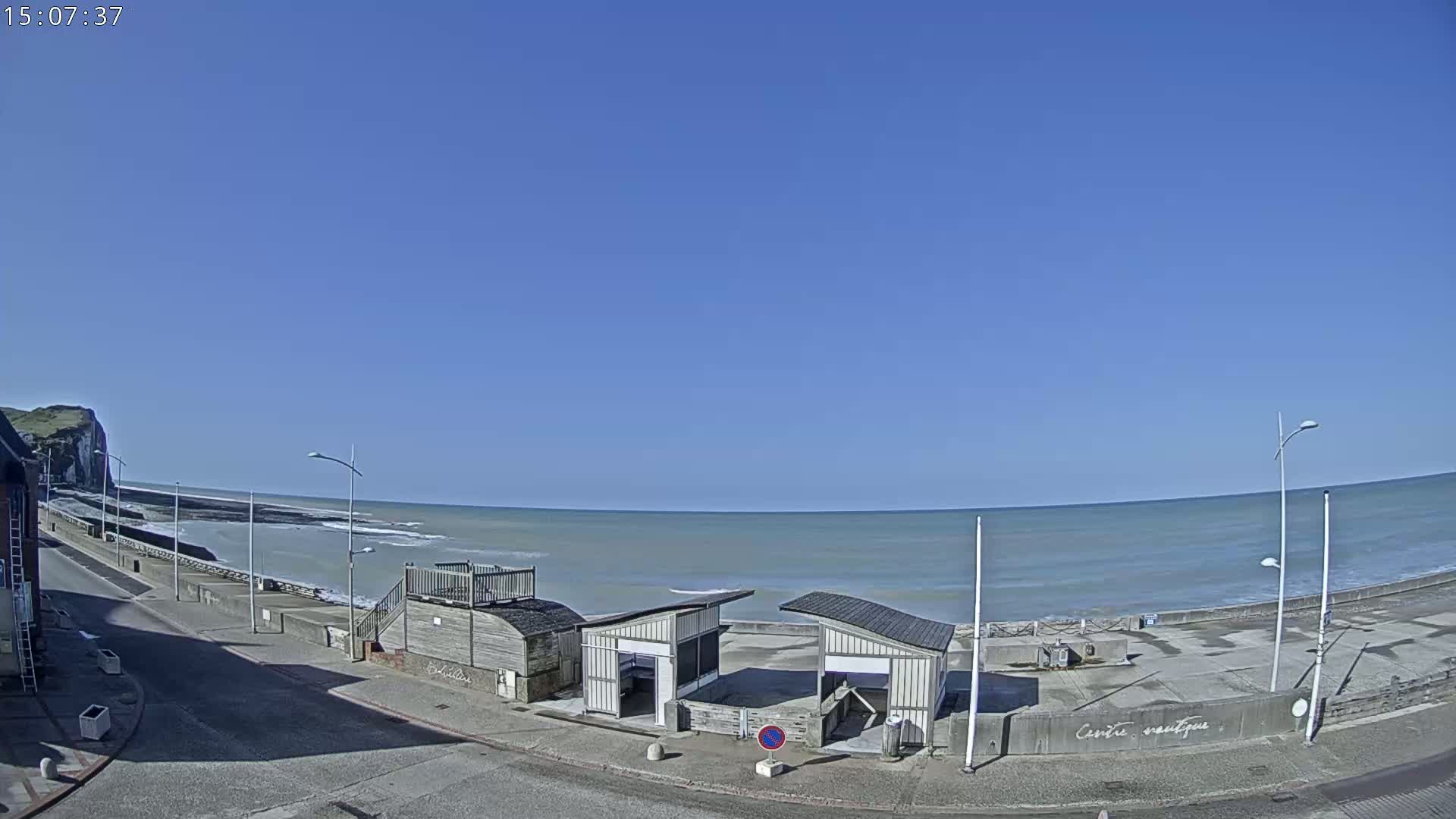 A seaside town on a clear, sunny day, shows a calm ocean, a paved beachfront, and two small, light-colored beachside structures.